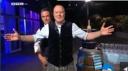 Zwei Männer in Tracht stehen vor einem Bierfass, einer breitet die Arme aus, im Hintergrund Gläser.
