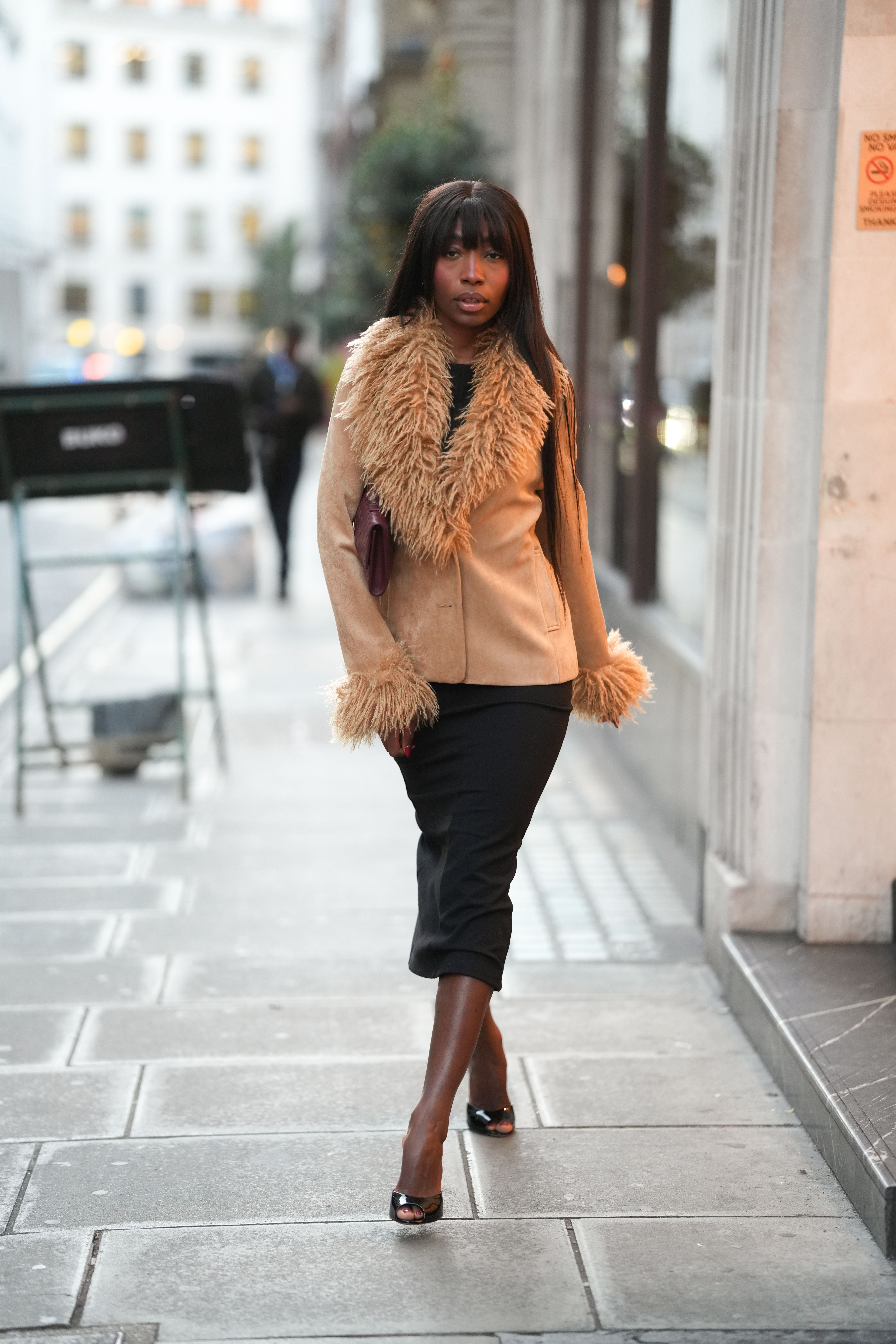 Frau mit beigem Shearling-Jacke und schwarzem Kleid läuft auf Gehweg in urbaner Umgebung.