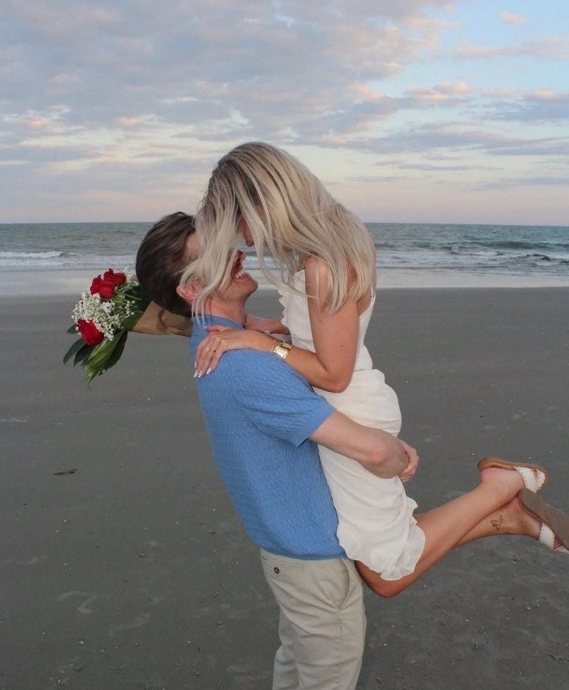Ein Mann hebt eine Frau am Strand hoch, sie hält einen Strauß roter Rosen.