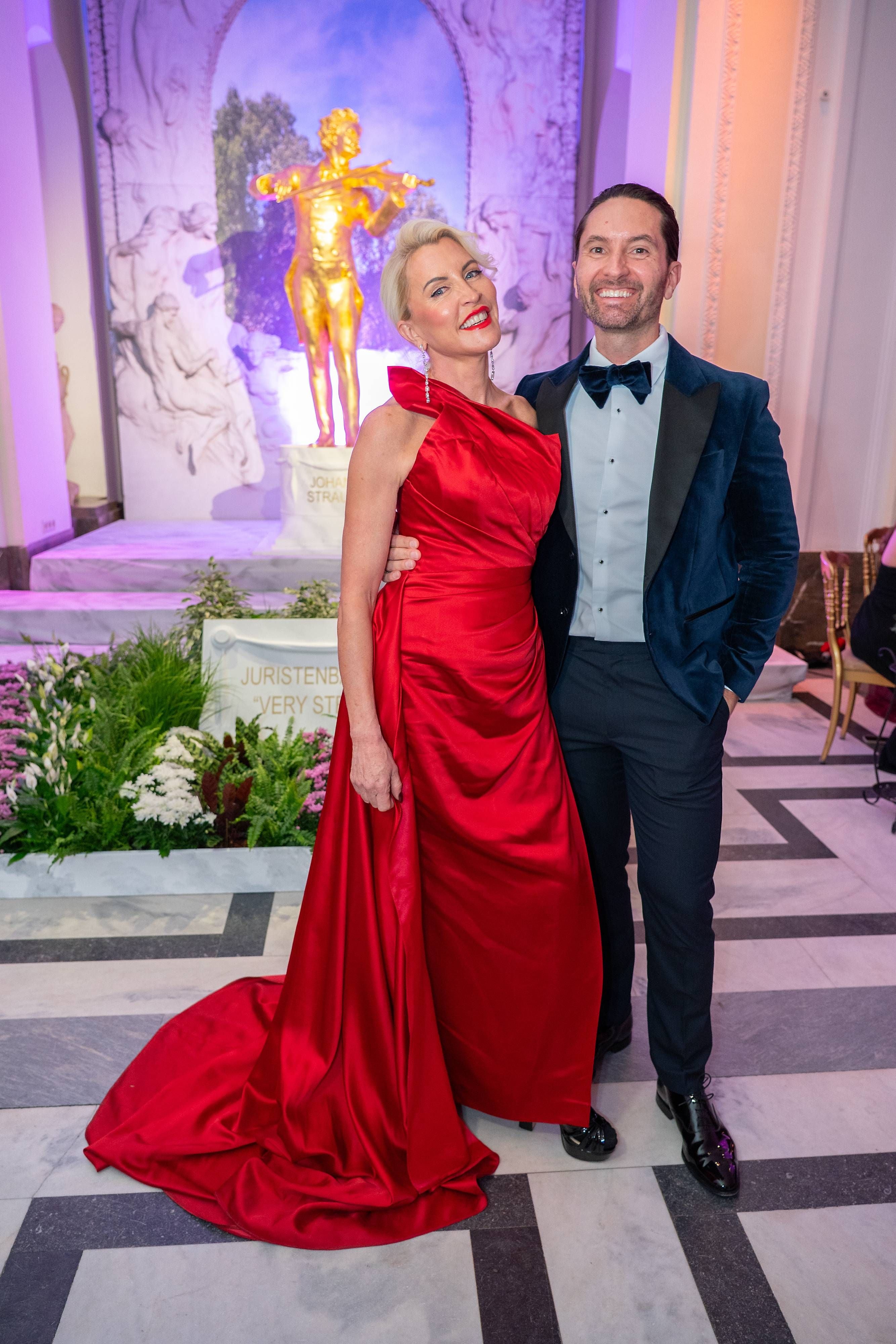 Ein Paar in Abendgarderobe posiert beim Juristenball 2025 vor einer goldenen Strauss-Statue.