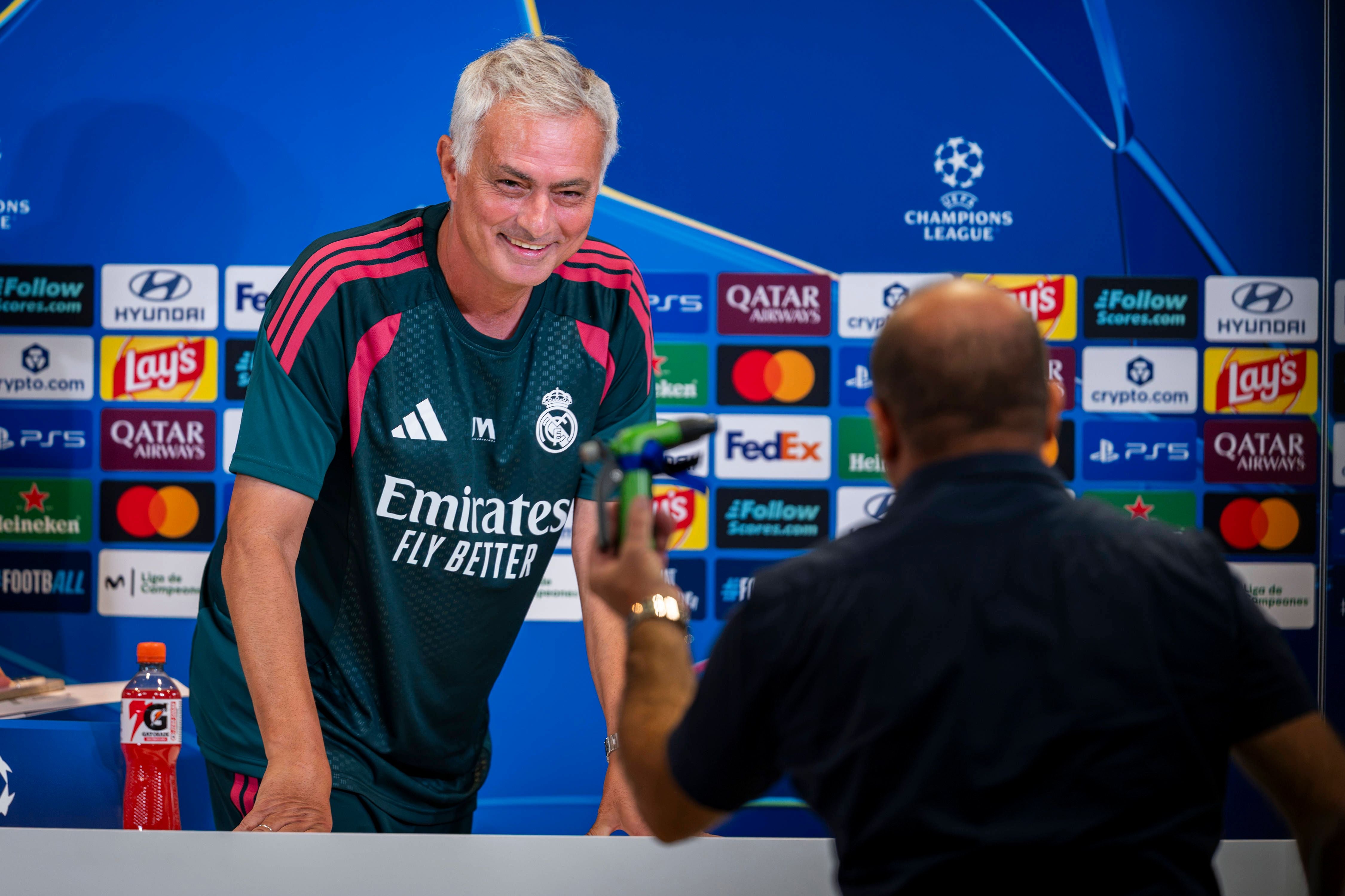 Jose Mourinho im dunklen Real-Madrid-Trainingsshirt lächelt bei einer Pressekonferenz vor einer Sponsorenwand.