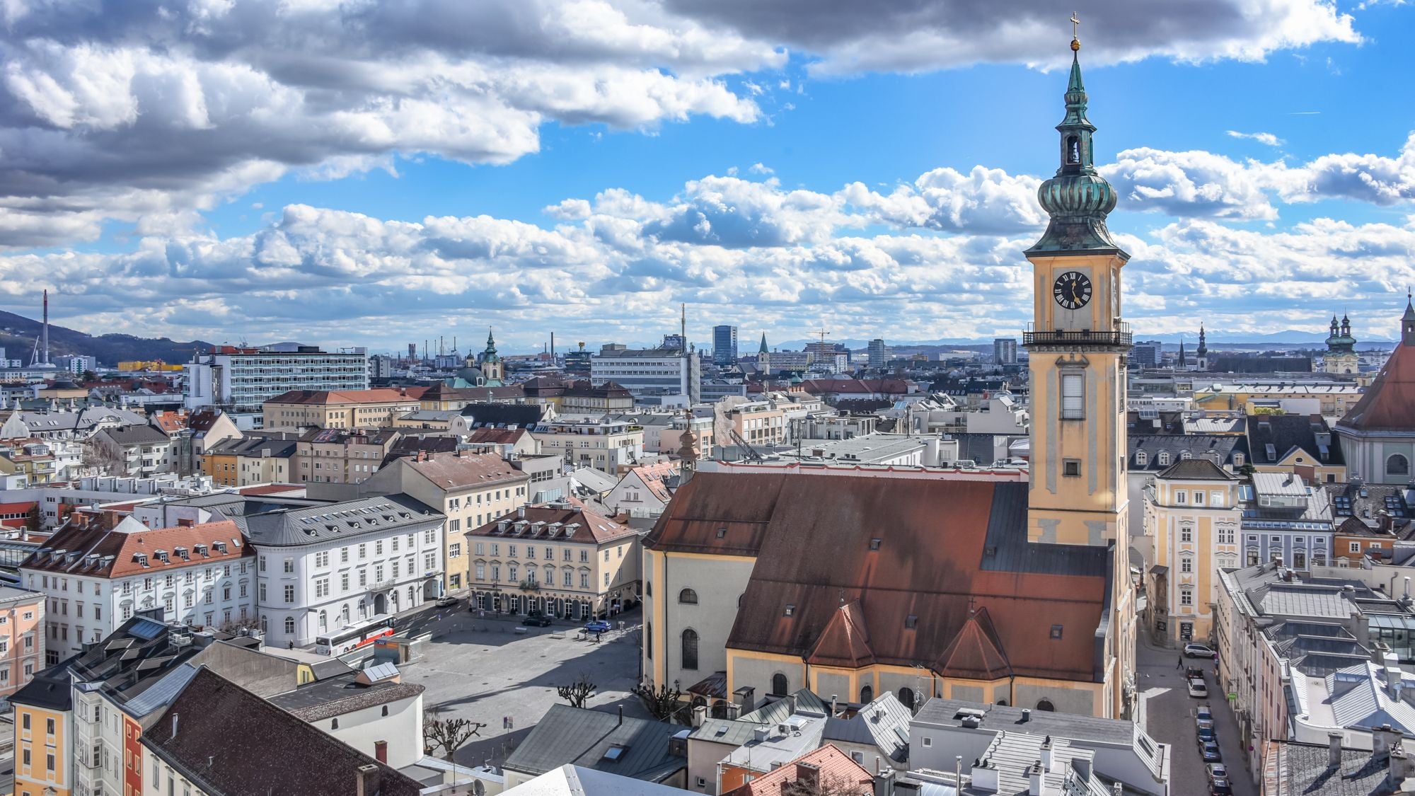 Stadtpanorama von Linz mit Wolkenhimmel und markantem Kirchturm im Vordergrund.