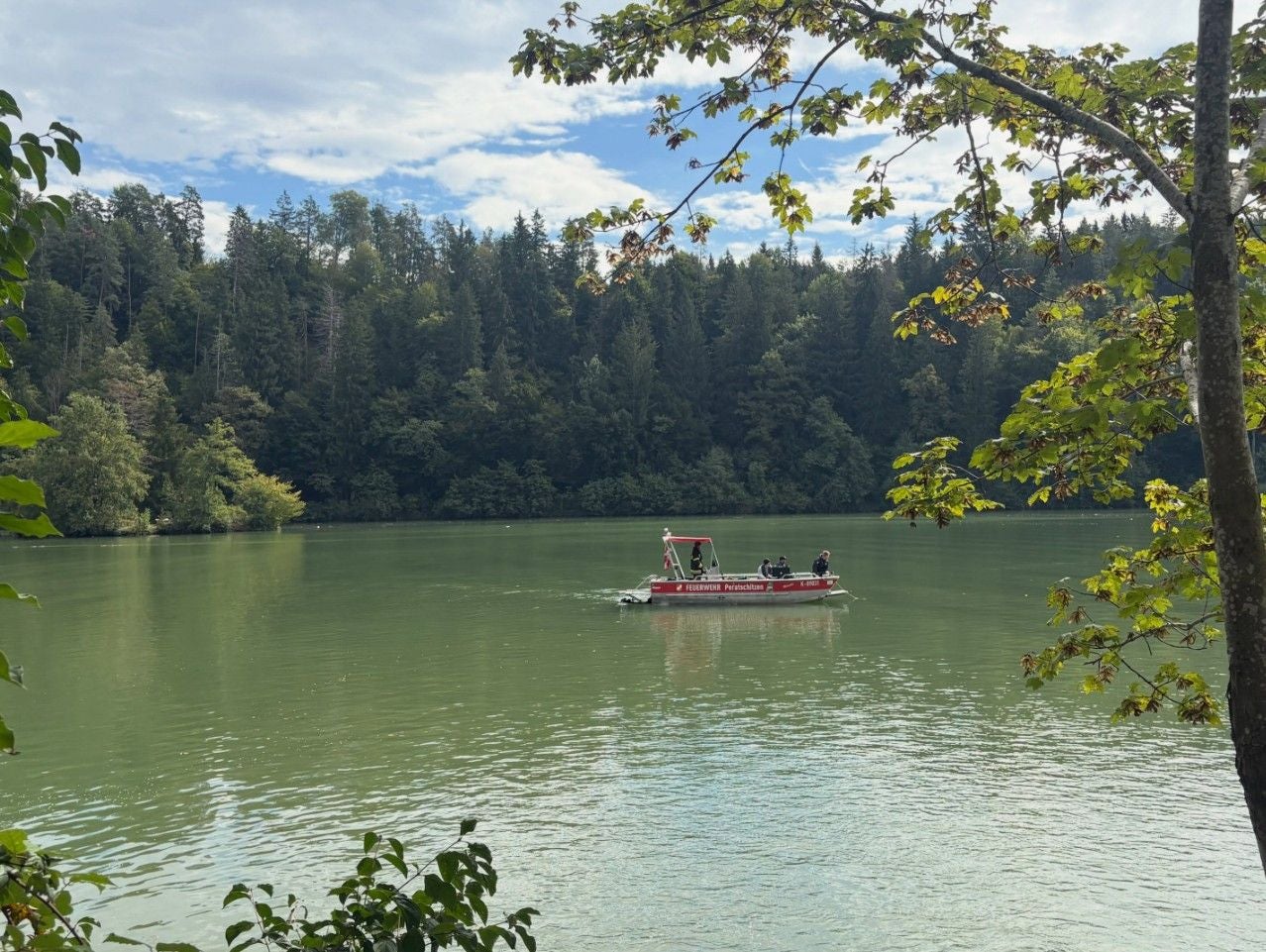 Feuerwehrboot mit Menschen auf einem See, umgeben von Bäumen, bei einer Suchaktion.