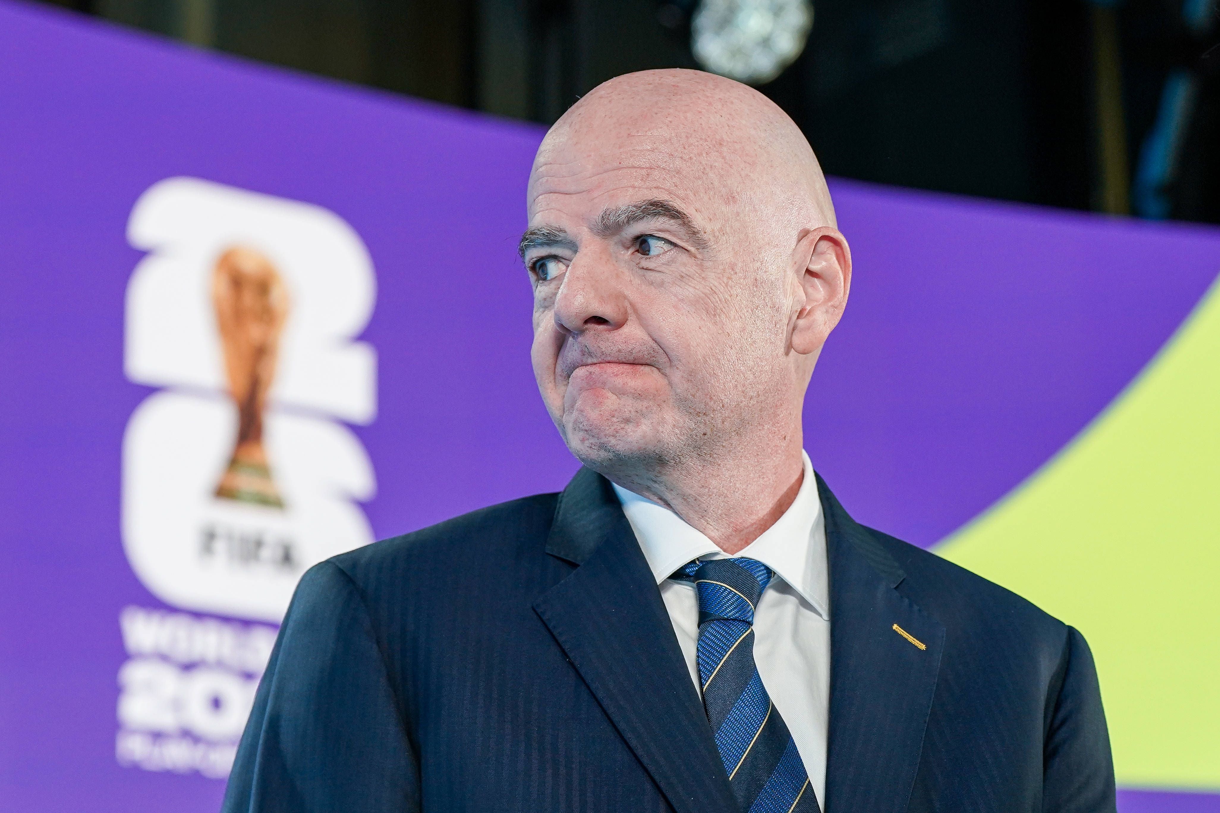 Ein glatzköpfiger Mann im Anzug steht vor einem lilafarbenen FIFA-WM-Hintergrund.