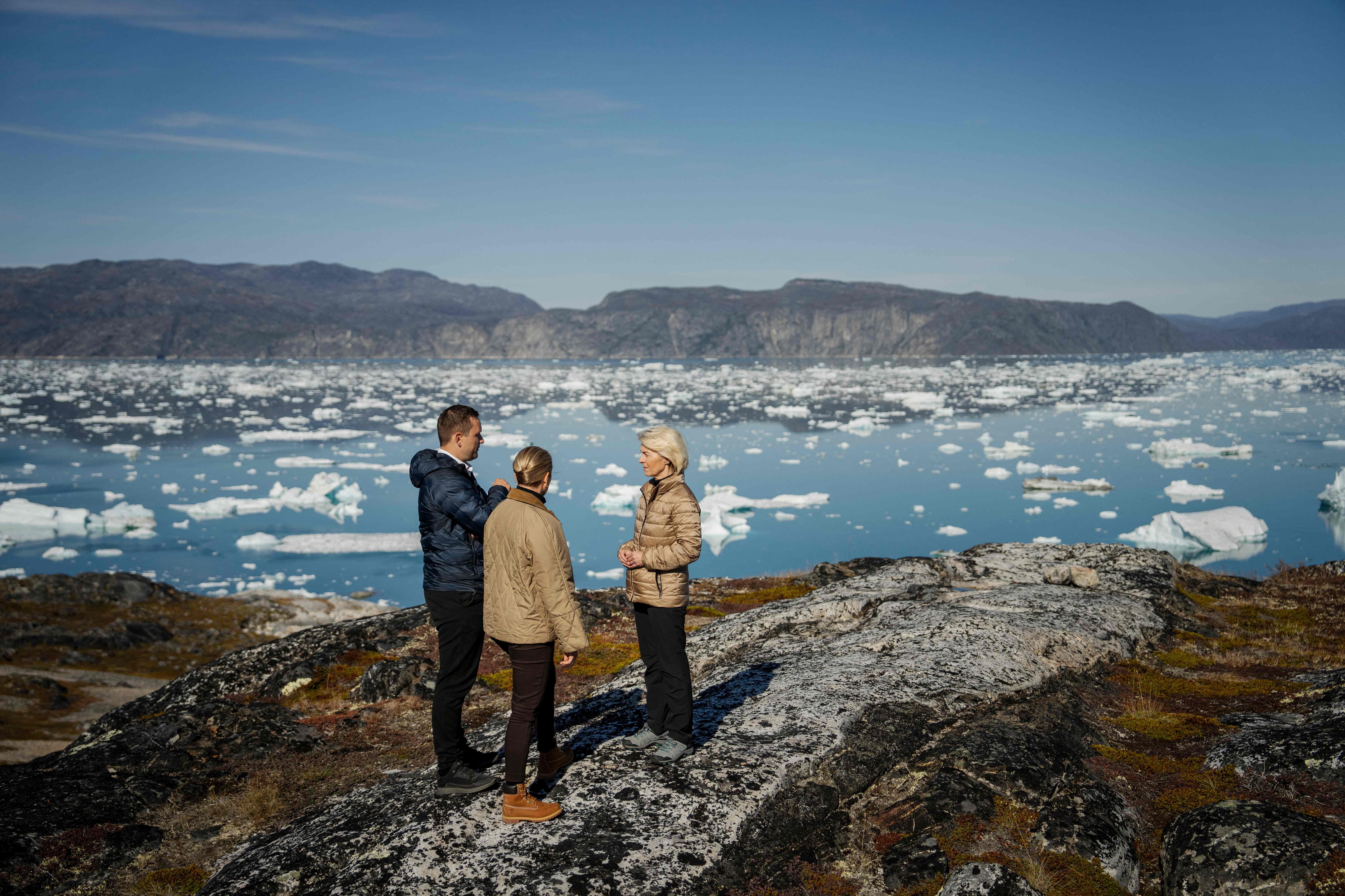 Drei Personen stehen auf Felsen und sprechen vor einem Meer mit Eisbergen und Bergen im Hintergrund.