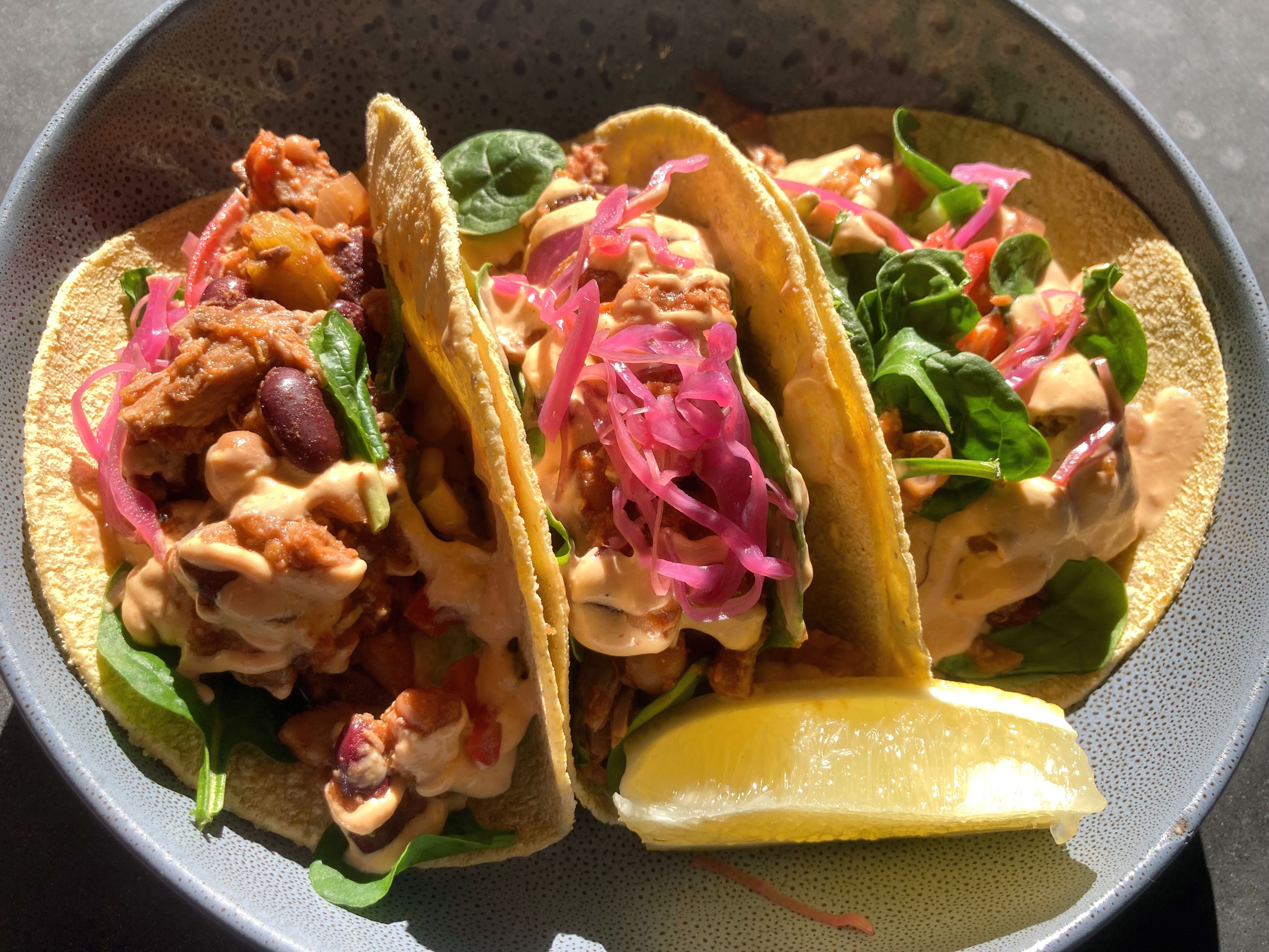 Drei Tacos mit Gemüse, Sauce, roten Bohnen, Spinat und Limettenschnitz auf einem Teller.