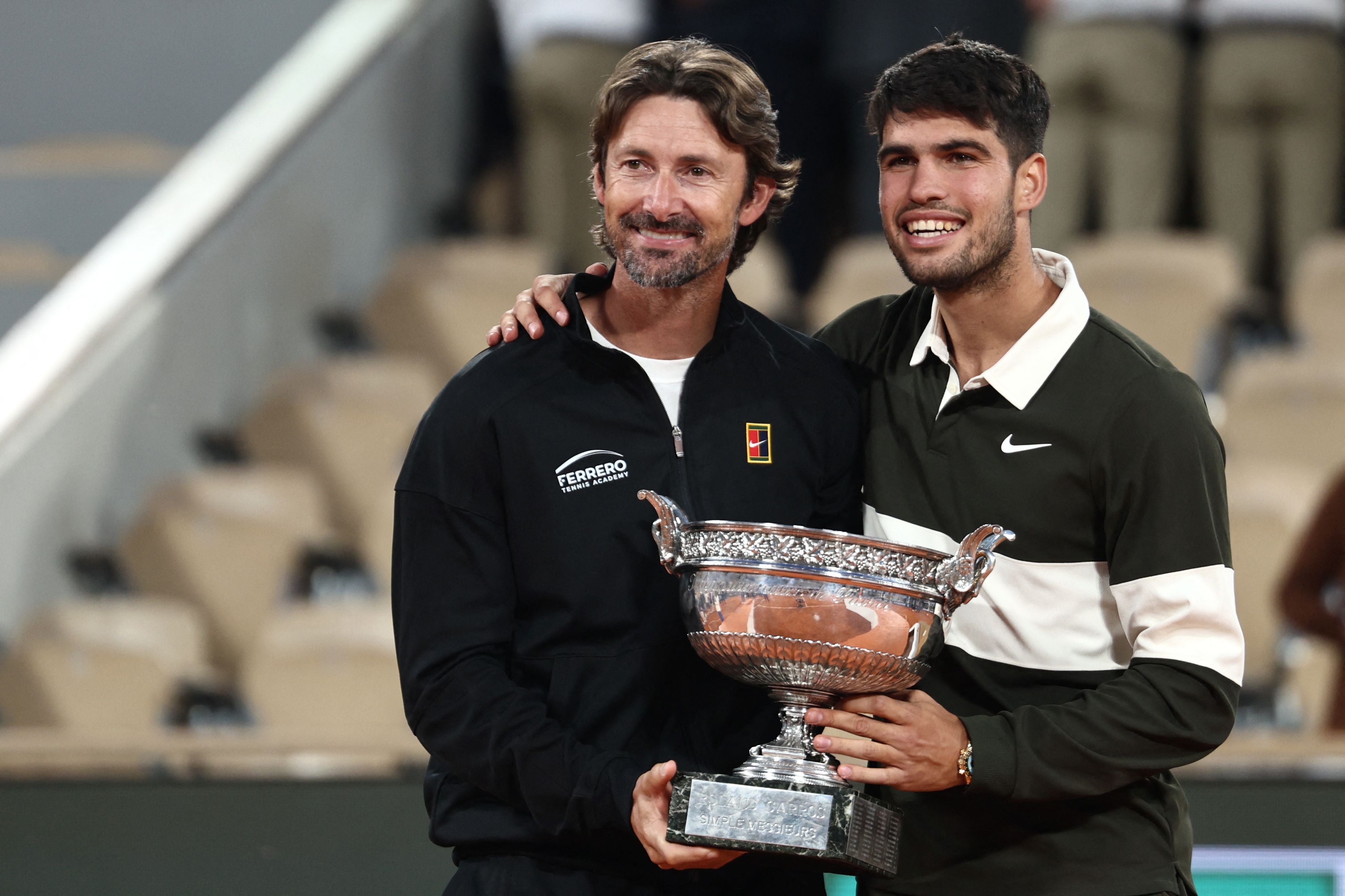 Carlos Alcaraz und sein Trainer posieren lächelnd mit dem French Open Pokal auf dem Tennisplatz.