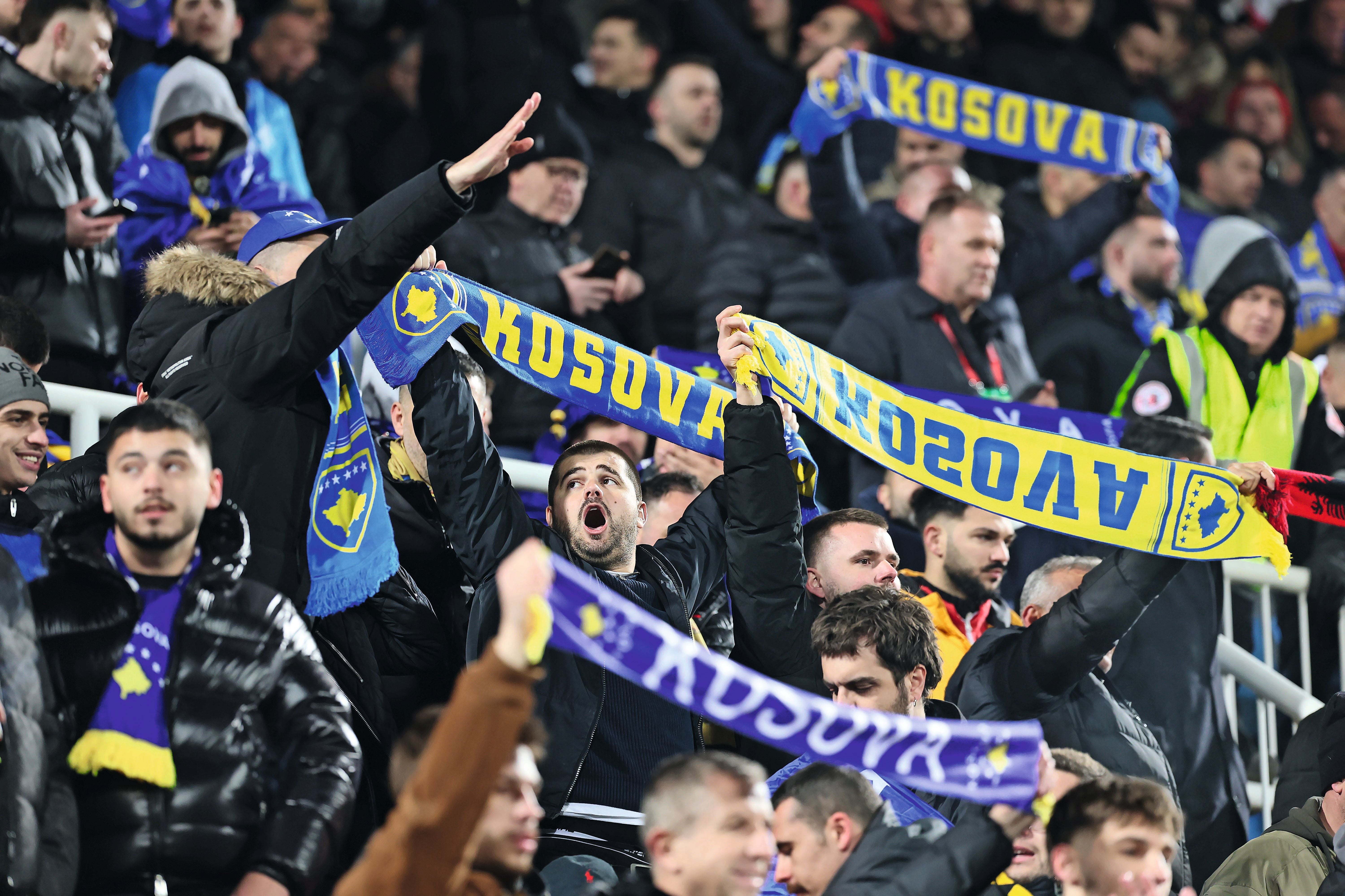 Anhänger der kosovarischen Nationalmannschaft jubeln im Stadion und halten gelb-blaue Fanschals mit der Aufschrift Kosova hoch.