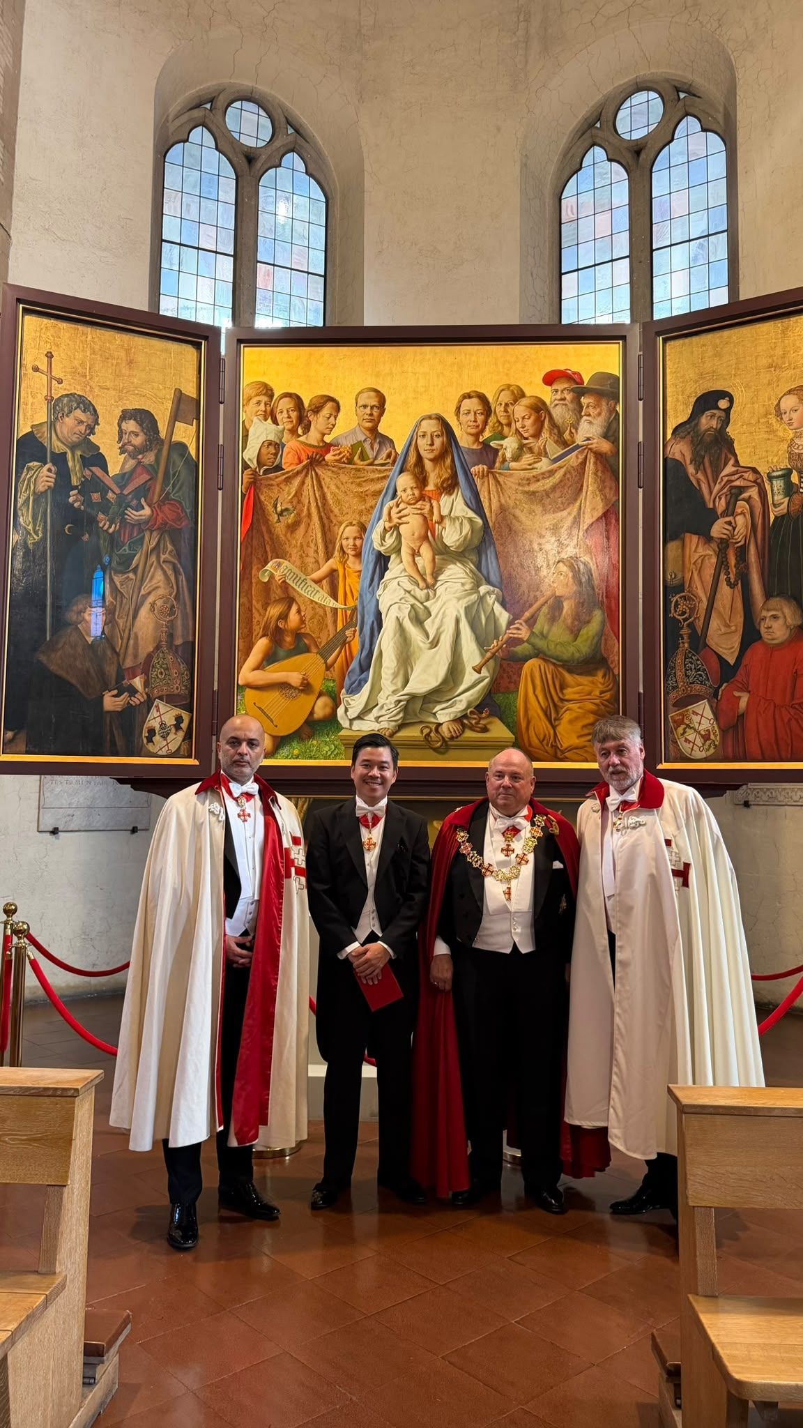 Vier Männer in festlicher Kleidung stehen vor einem Altarbild in einer Kirche mit hohen Fenstern.