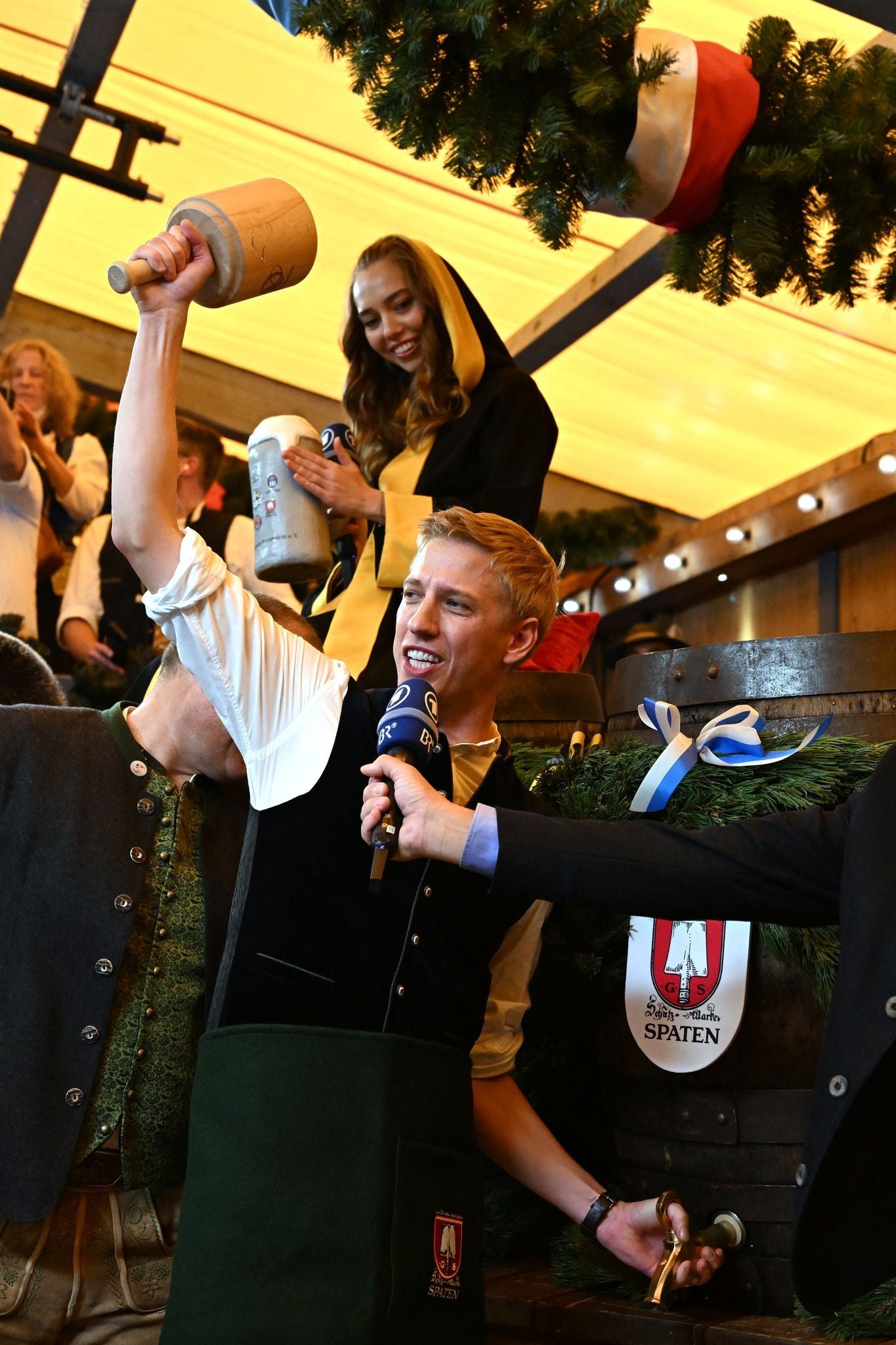 Ein Mann schlägt mit einem Holzhammer ein Bierfass an, umgeben von festlich gekleideten Menschen.