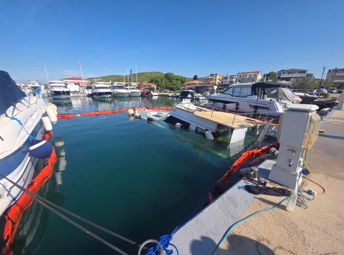 Halb gesunkene Jacht im Hafen, umgeben von Absperrungen und weiteren Booten bei klarem Wetter.
