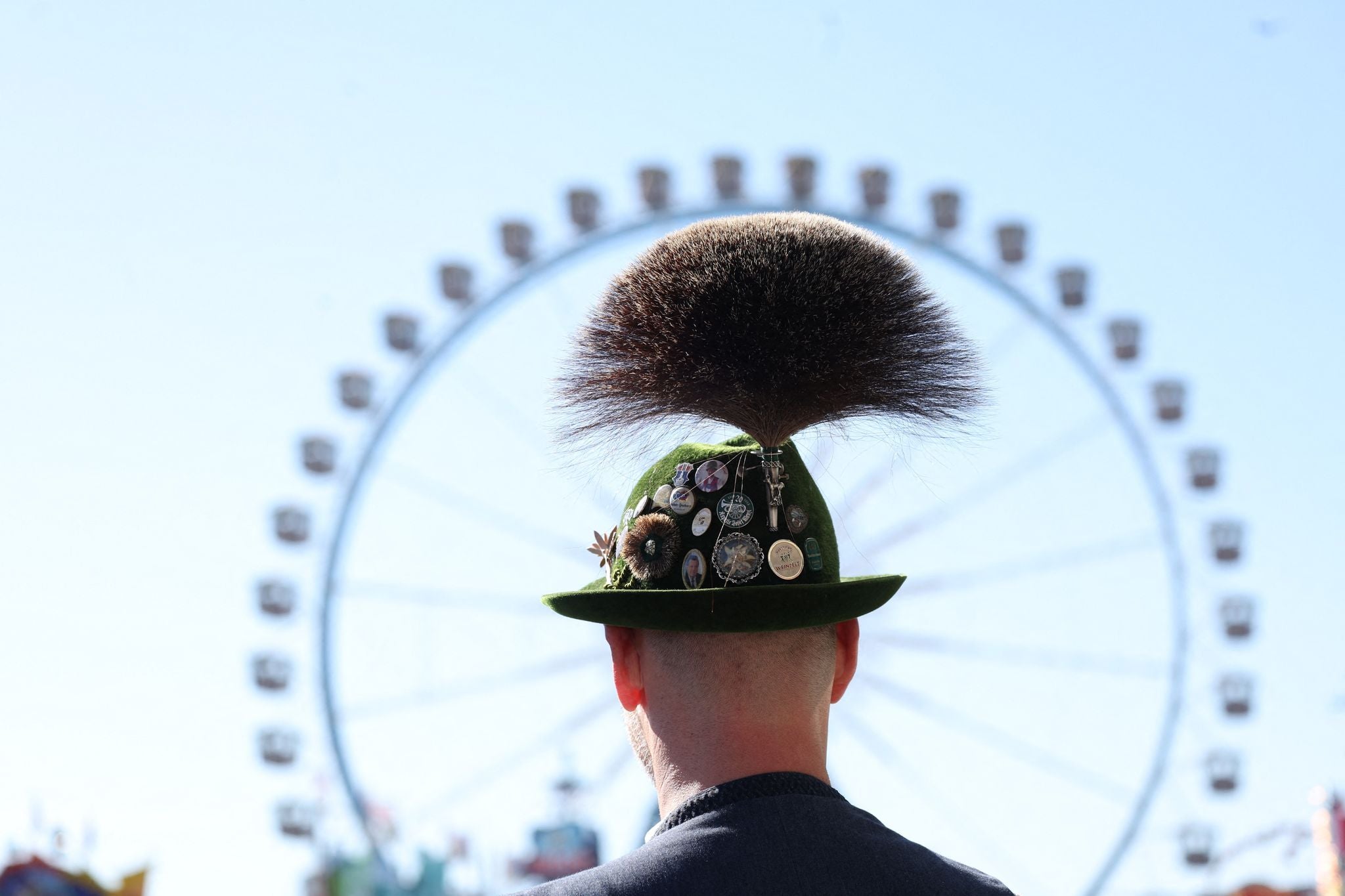 Ein Mann in Tracht mit Gamsbart-Hut steht vor einem Riesenrad auf dem Oktoberfest.
