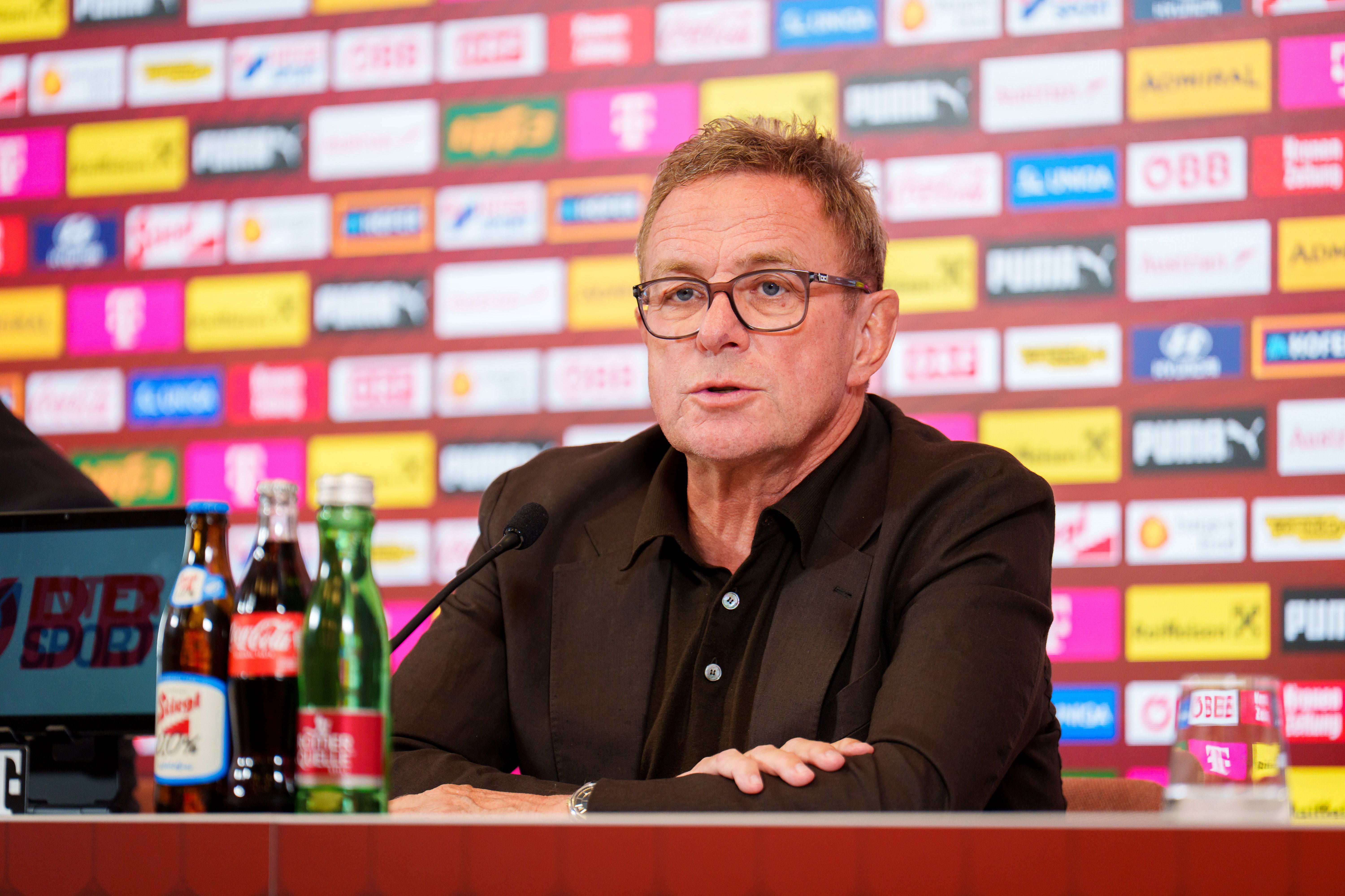 Ein Mann spricht bei einer Pressekonferenz vor einer Sponsorenwand und mehreren Getränken.