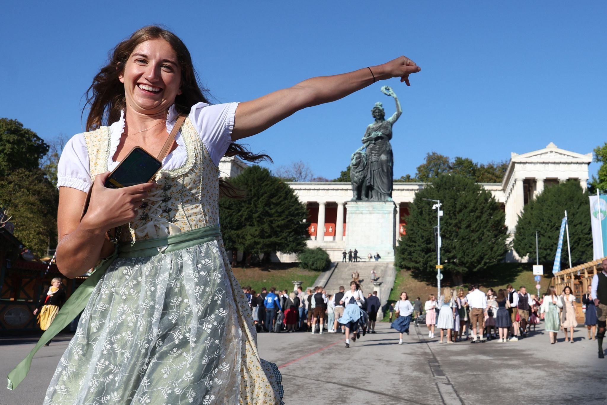 Junge Frau in Dirndl lacht vor der Bavaria-Statue beim Oktoberfest in München, viele Menschen im Hintergrund.