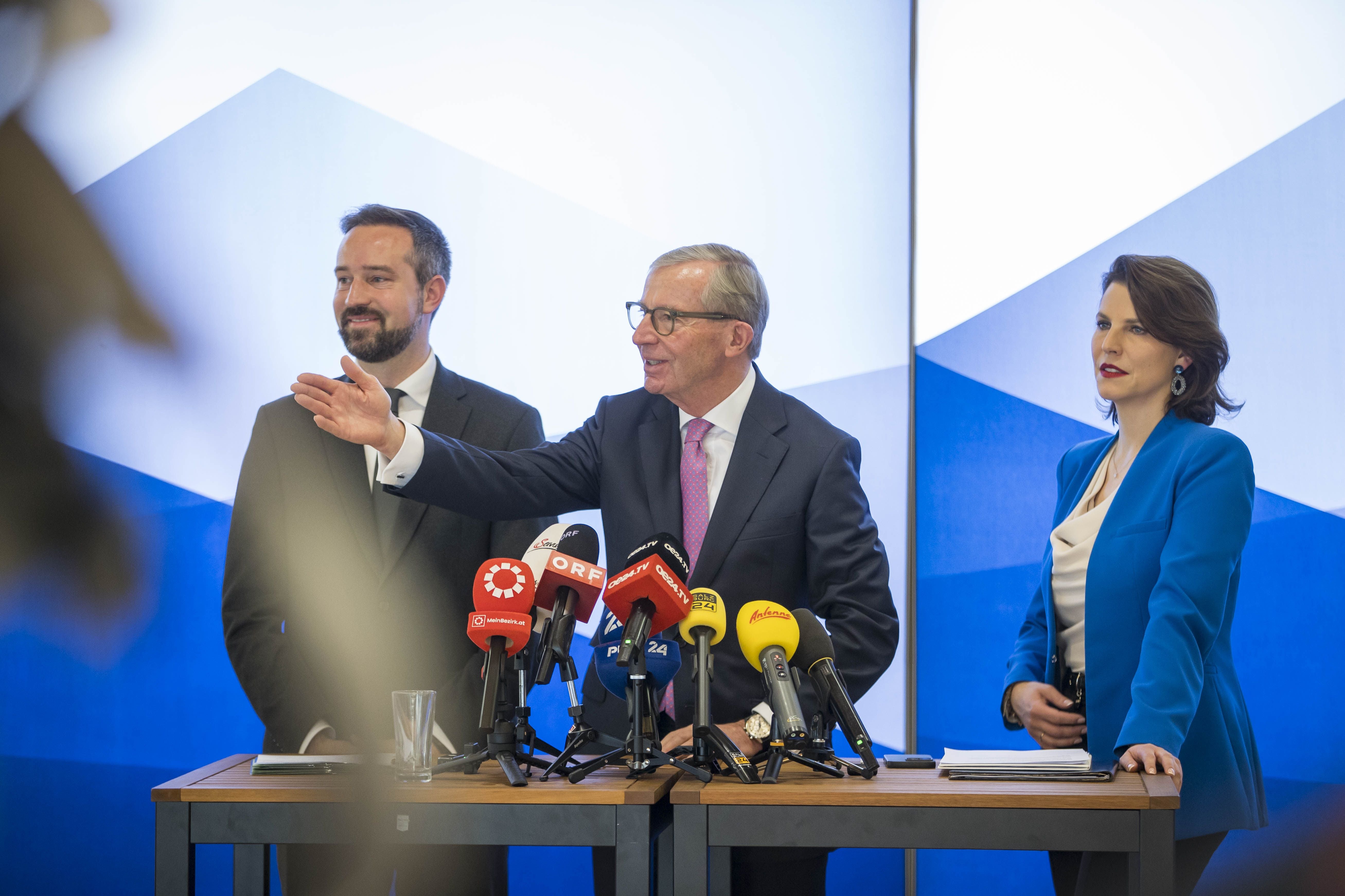 Drei Personen im Anzug bei einer Pressekonferenz mit mehreren Mikrofonen vor blauem Hintergrund.