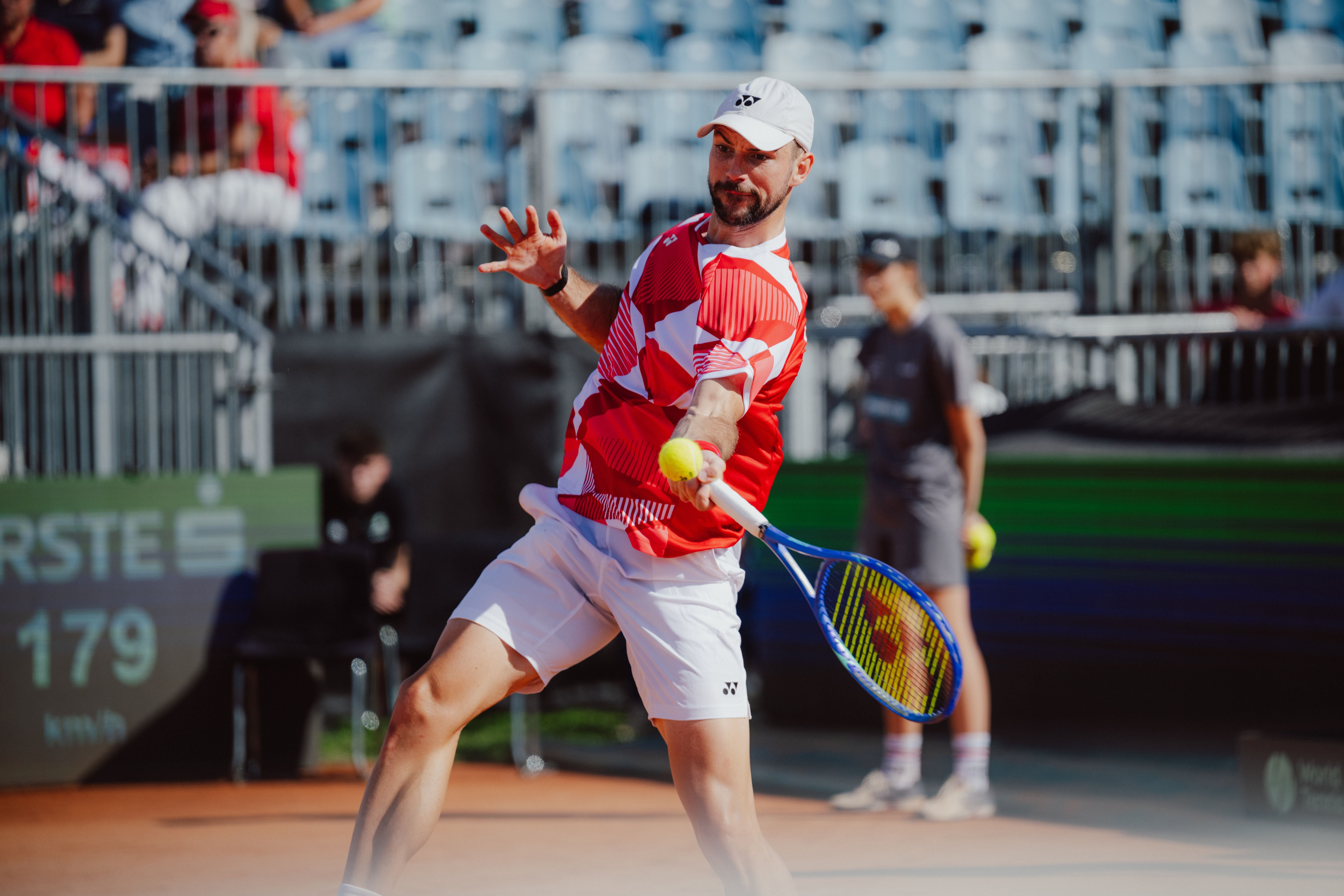 Ein Tennisspieler im roten Trikot schlägt auf einem Sandplatz den Ball mit einem Schläger.