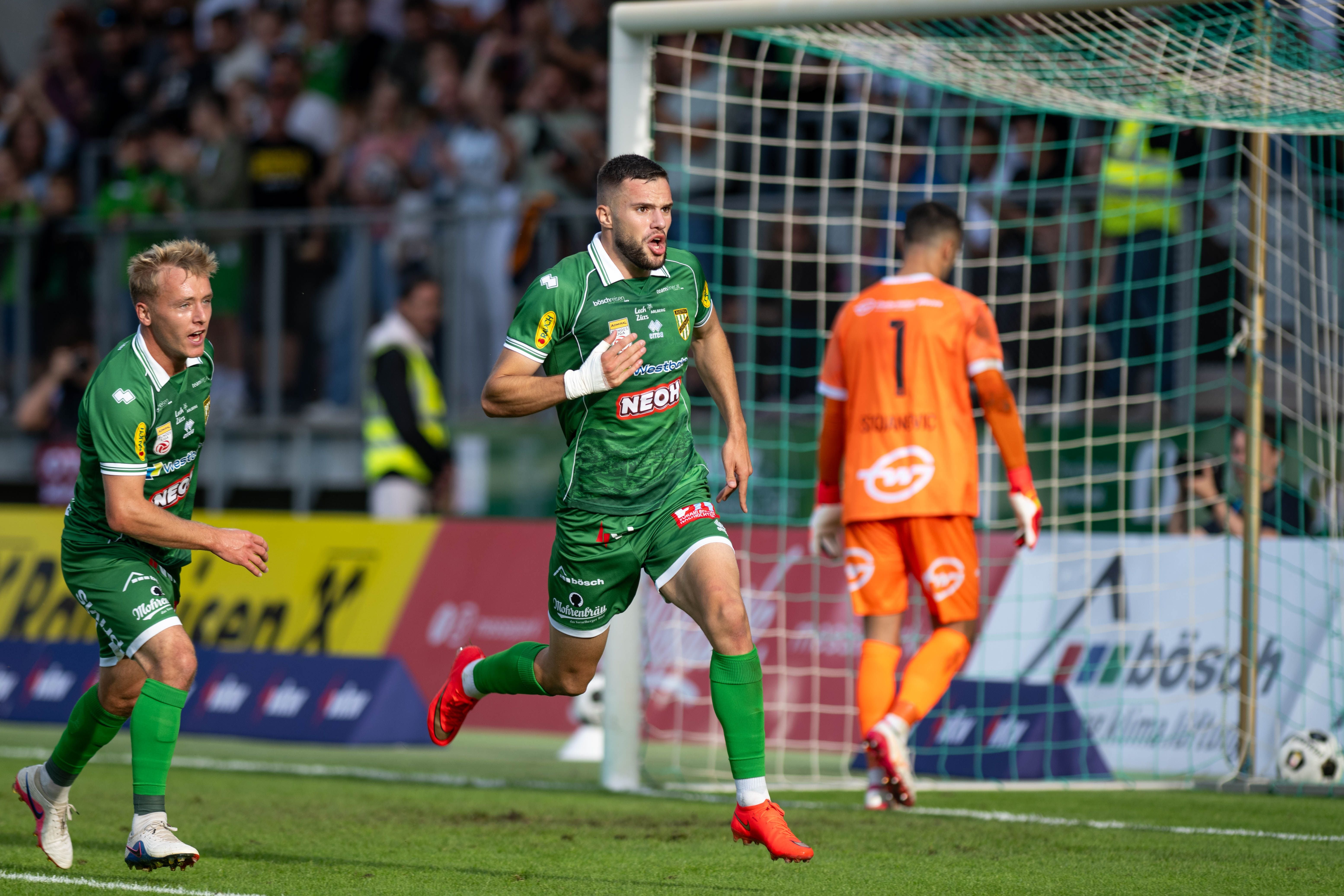 Zwei Fußballspieler jubeln, während ein enttäuschter Torwart im Hintergrund steht.