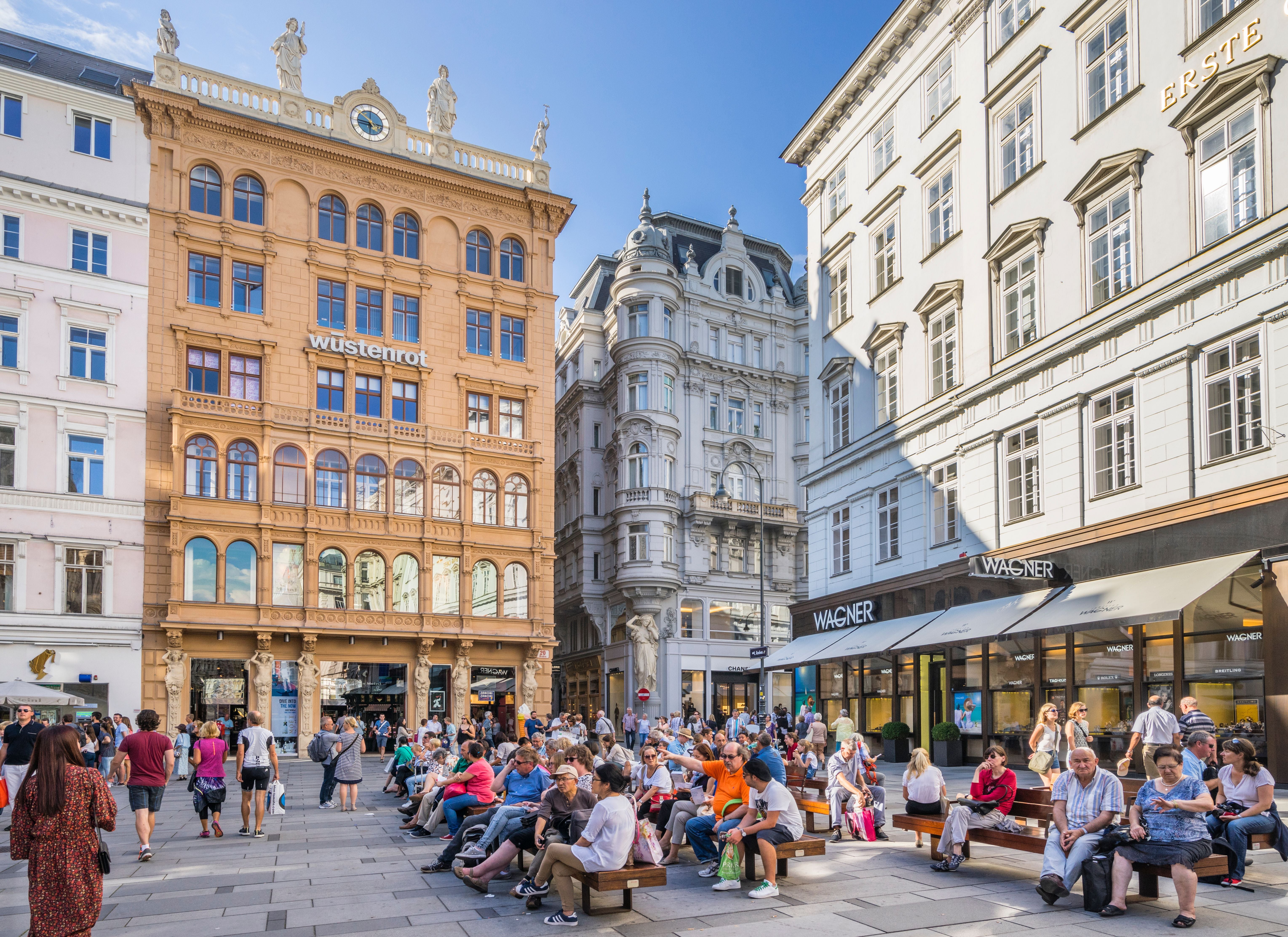 Viele Menschen sitzen auf Bänken in der Fußgängerzone der Grabenstraße in Wien bei Sonnenschein.