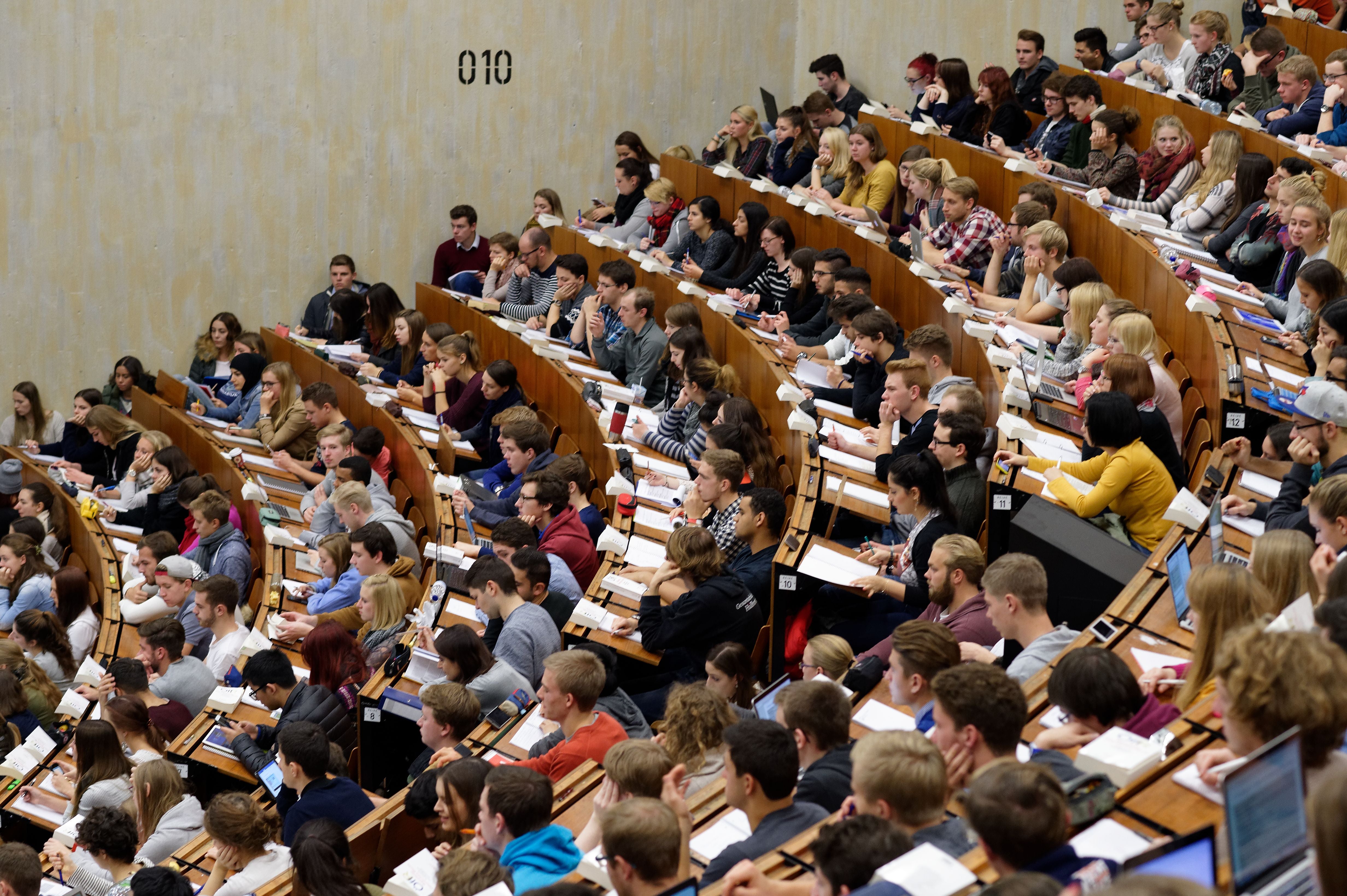 Viele Studierende sitzen im Hörsaal und hören einer Vorlesung zu, einige machen Notizen.