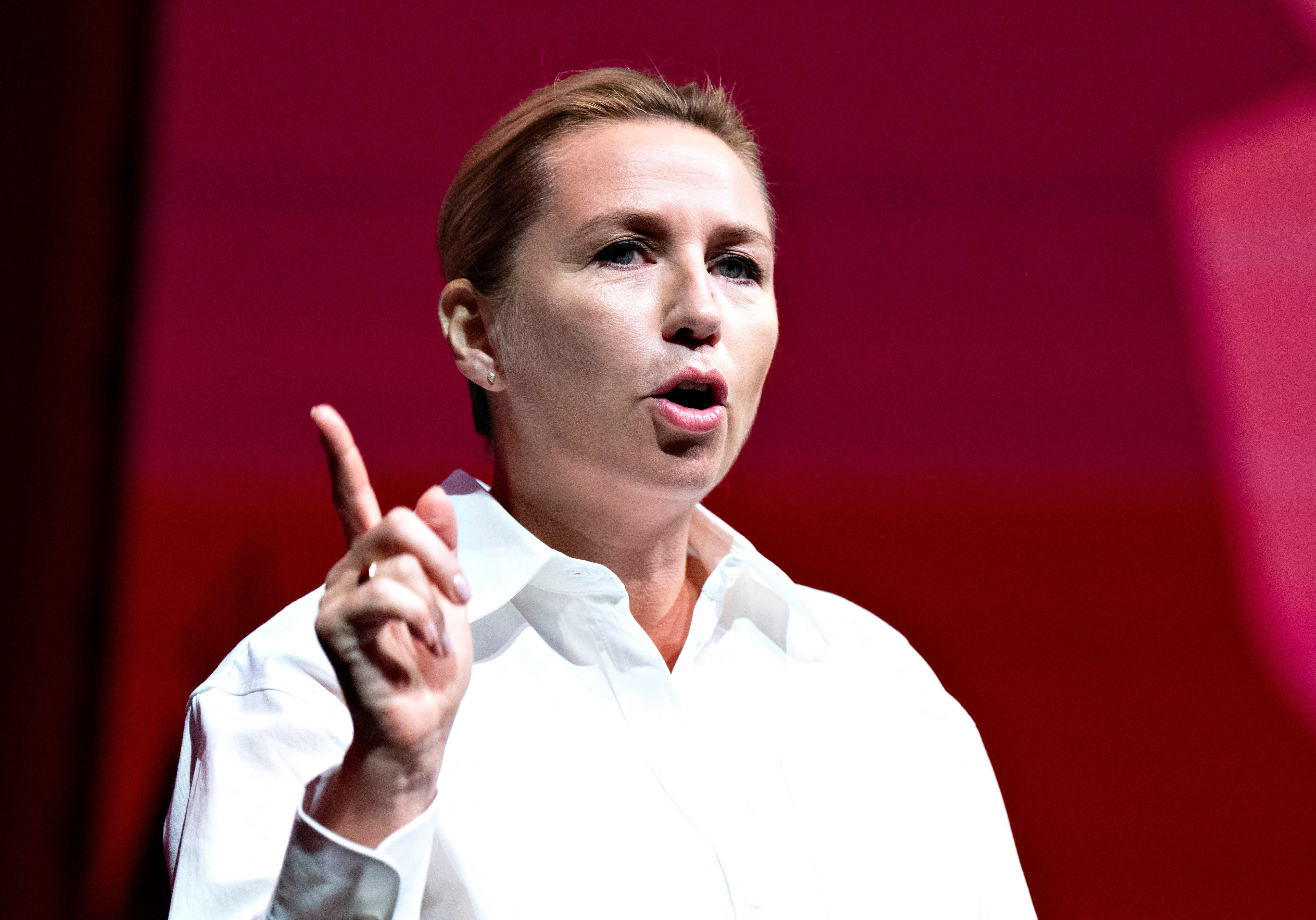Frau in weißem Hemd spricht und hebt einen Finger, vor rotem Hintergrund.