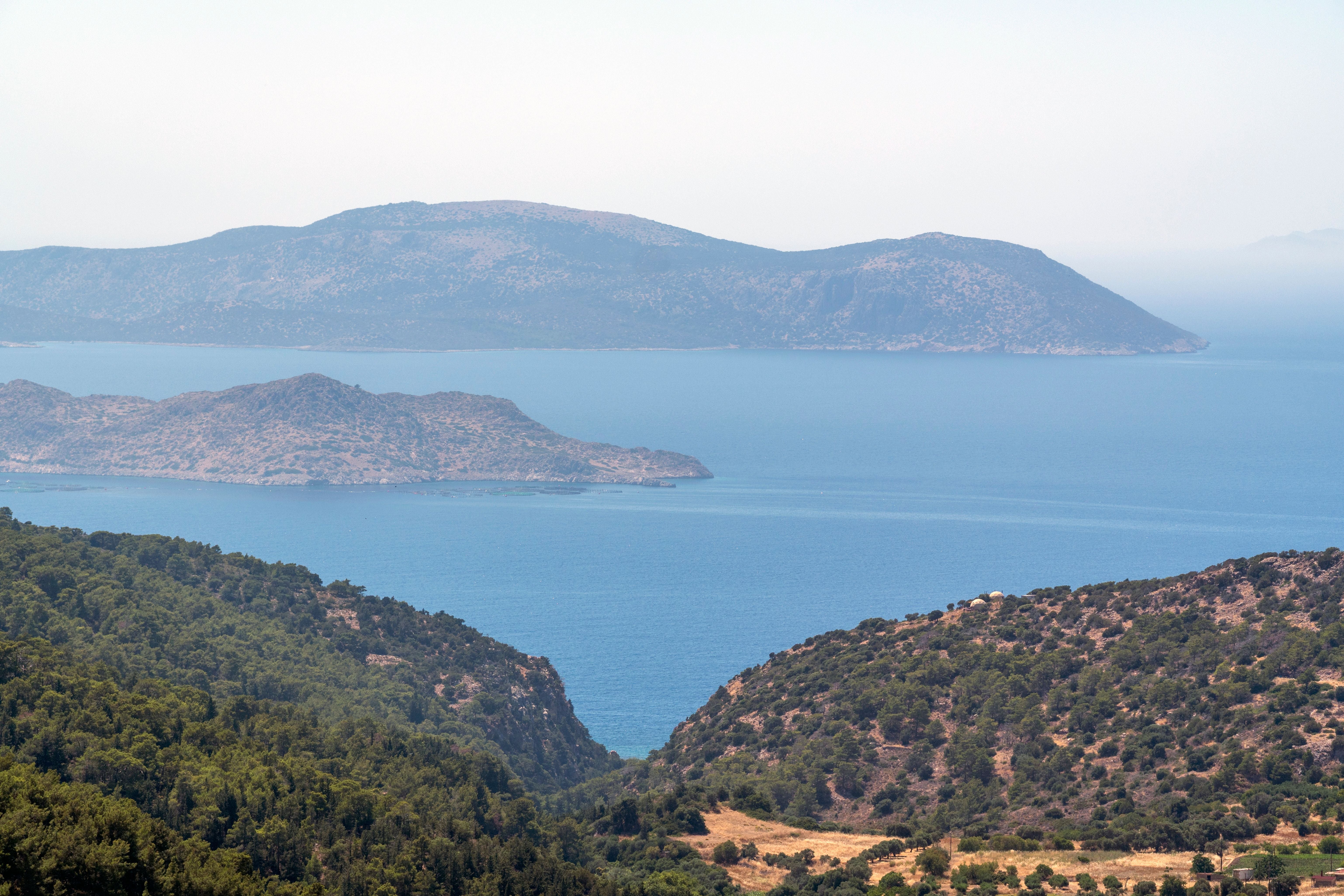 Grüne Hügel auf Rhodos überblicken das blaue Meer, dahinter liegen Inseln unter klarem Himmel.