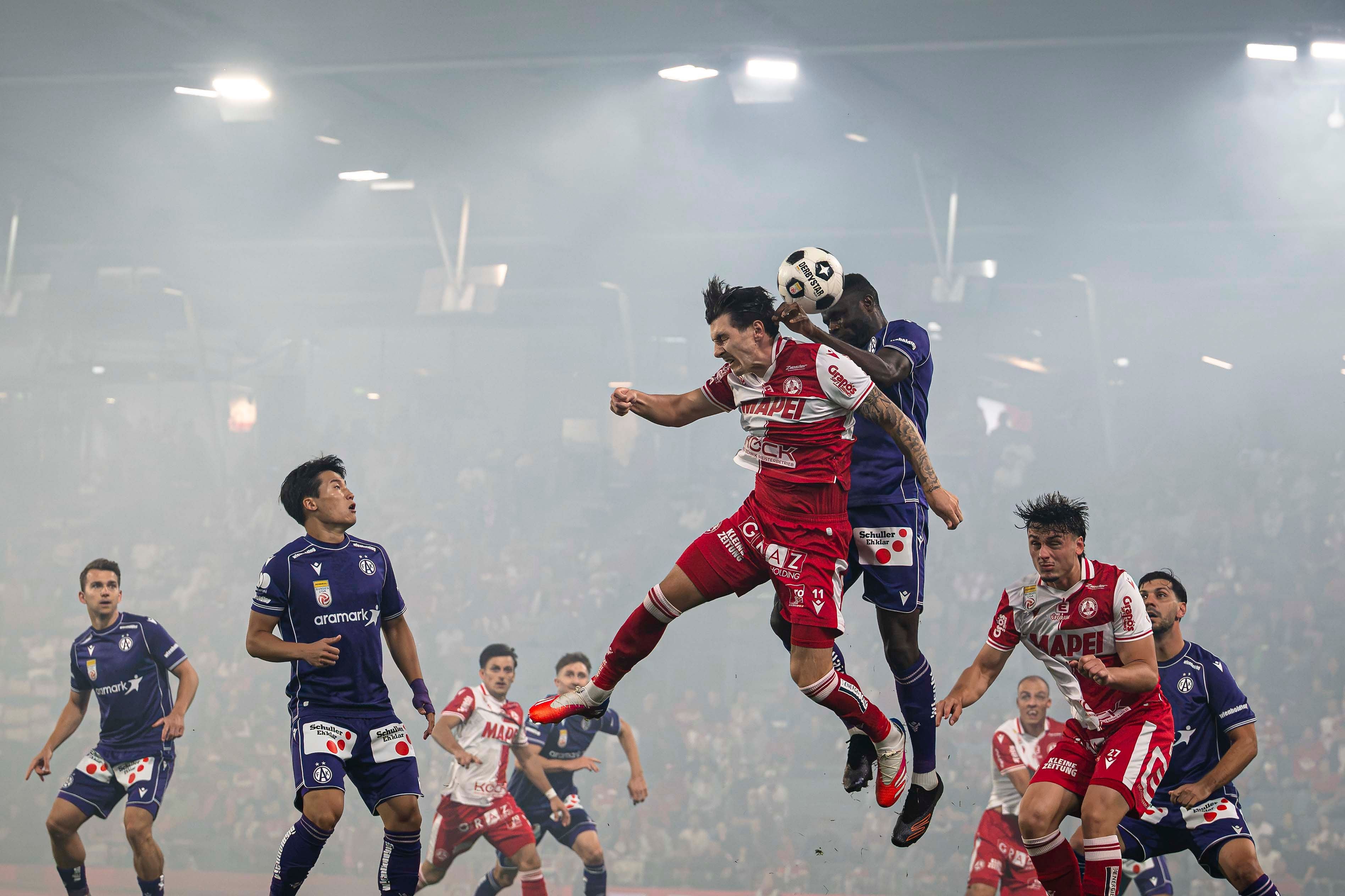 Fußballspieler springen zum Kopfball in einem nebligen Stadion während eines Spiels.