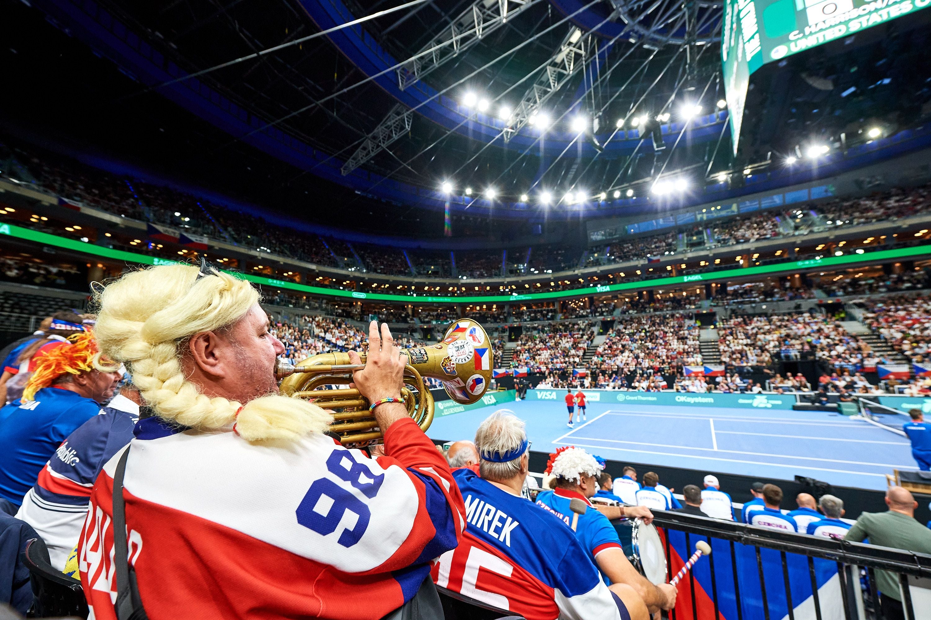 Fans im Kostüm feuern mit Instrumenten ein Tennis-Doppelspiel in einer vollen Arena an.