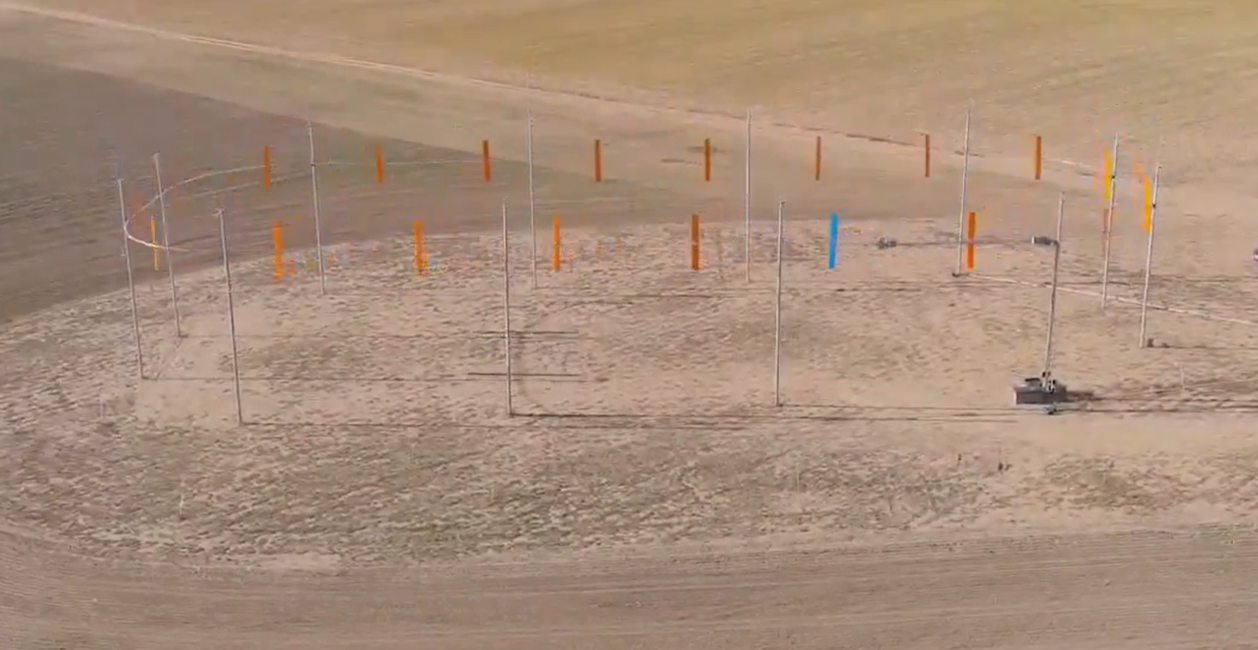 Mehrere Messmasten mit bunten Markierungen stehen auf einem sandigen Feld zur Windkraftmessung.