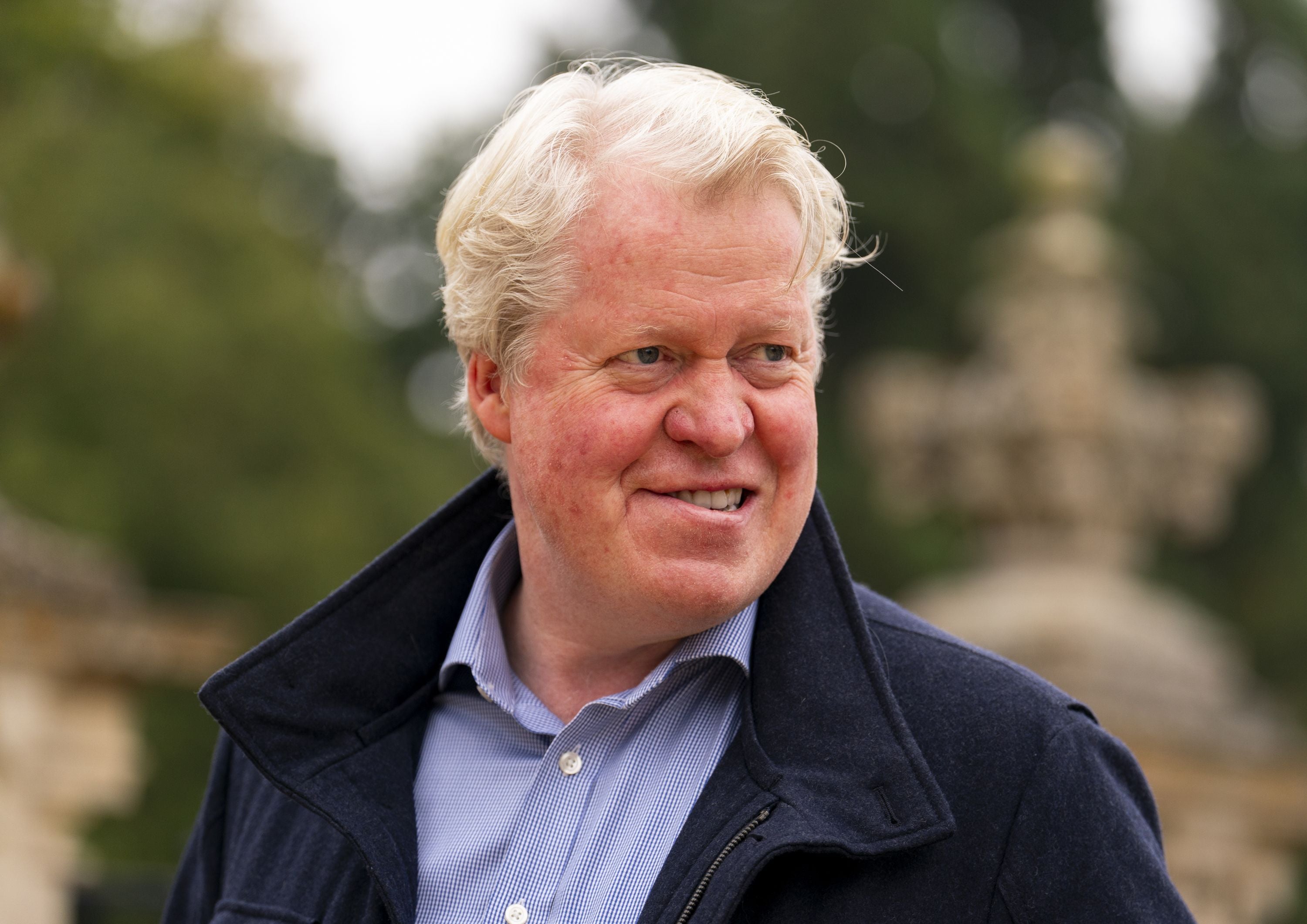 Ein Mann mit blondem Haar lächelt im Freien und trägt Hemd und dunkle Jacke.