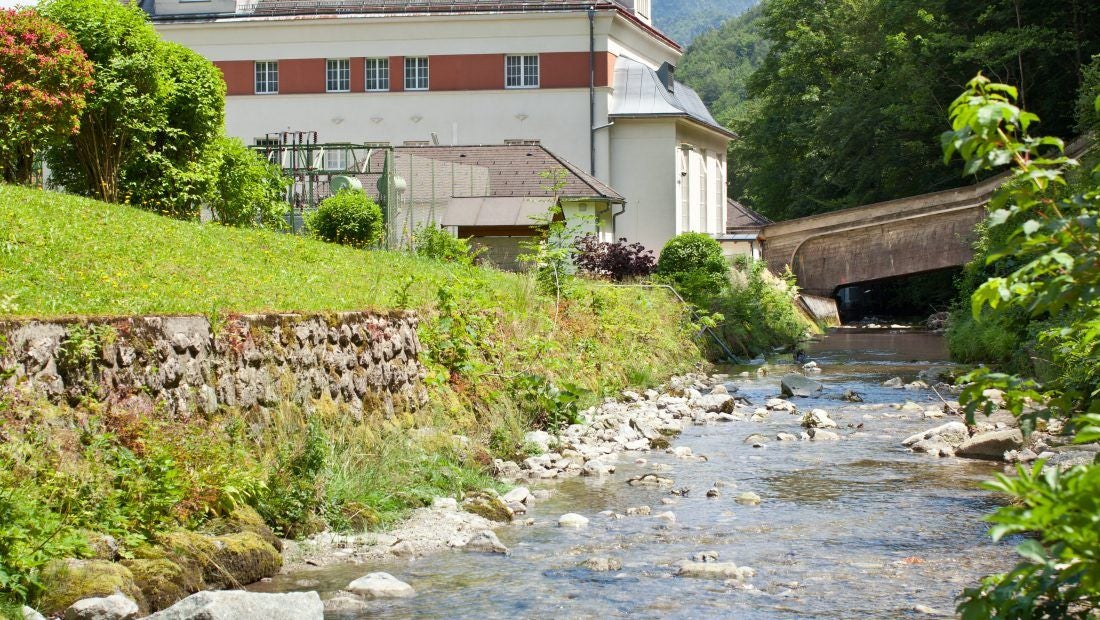 Kleinwasserkraftwerk an einem Bach mit begrüntem Ufer und Gebäude im Hintergrund.