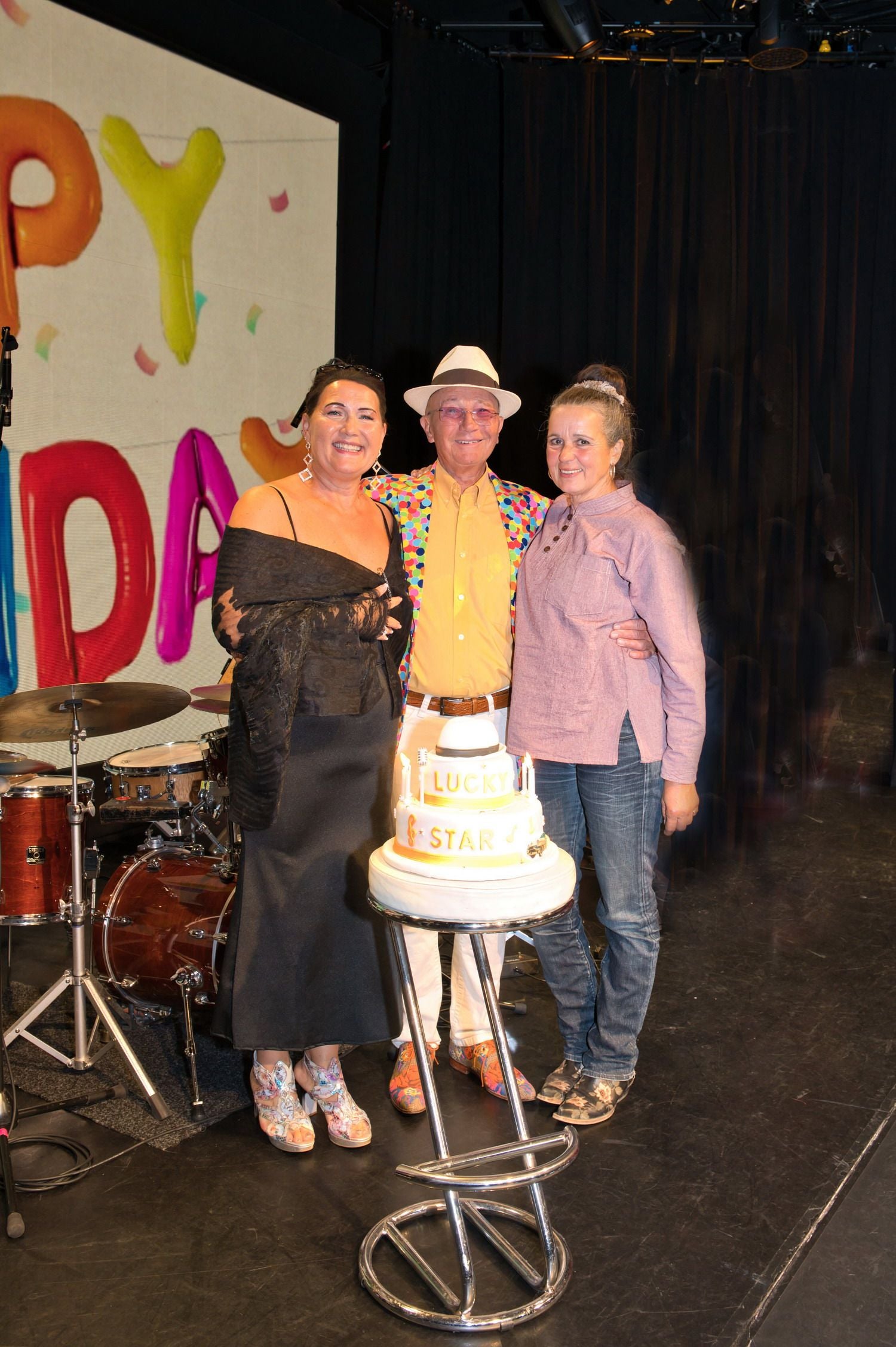 Drei Menschen stehen vor einer Geburtstagstorte auf einer Bühne mit Happy Birthday-Deko im Hintergrund.