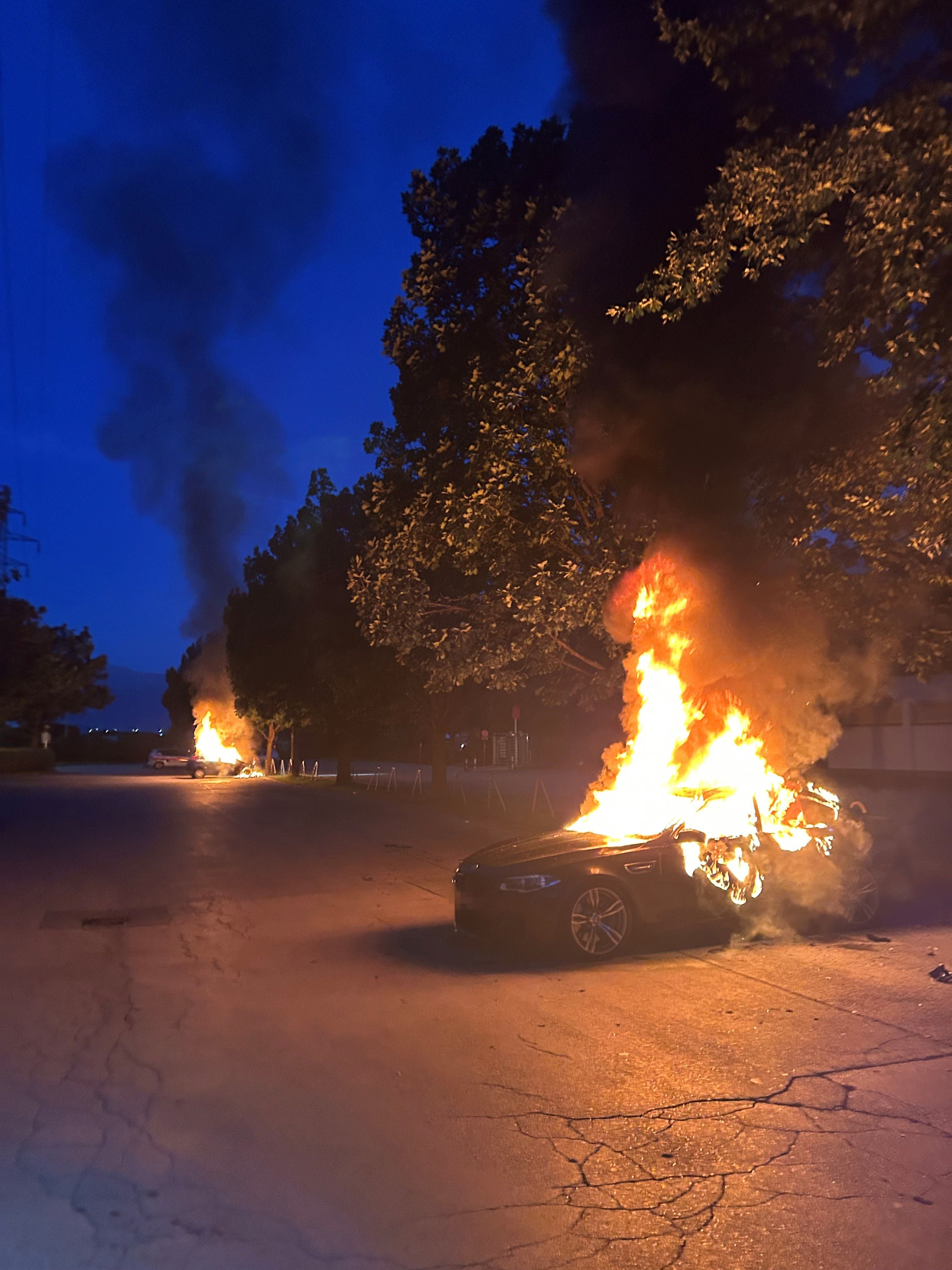Zwei Autos stehen nachts auf einem Parkplatz in Innsbruck in Flammen, dichter Rauch steigt auf.