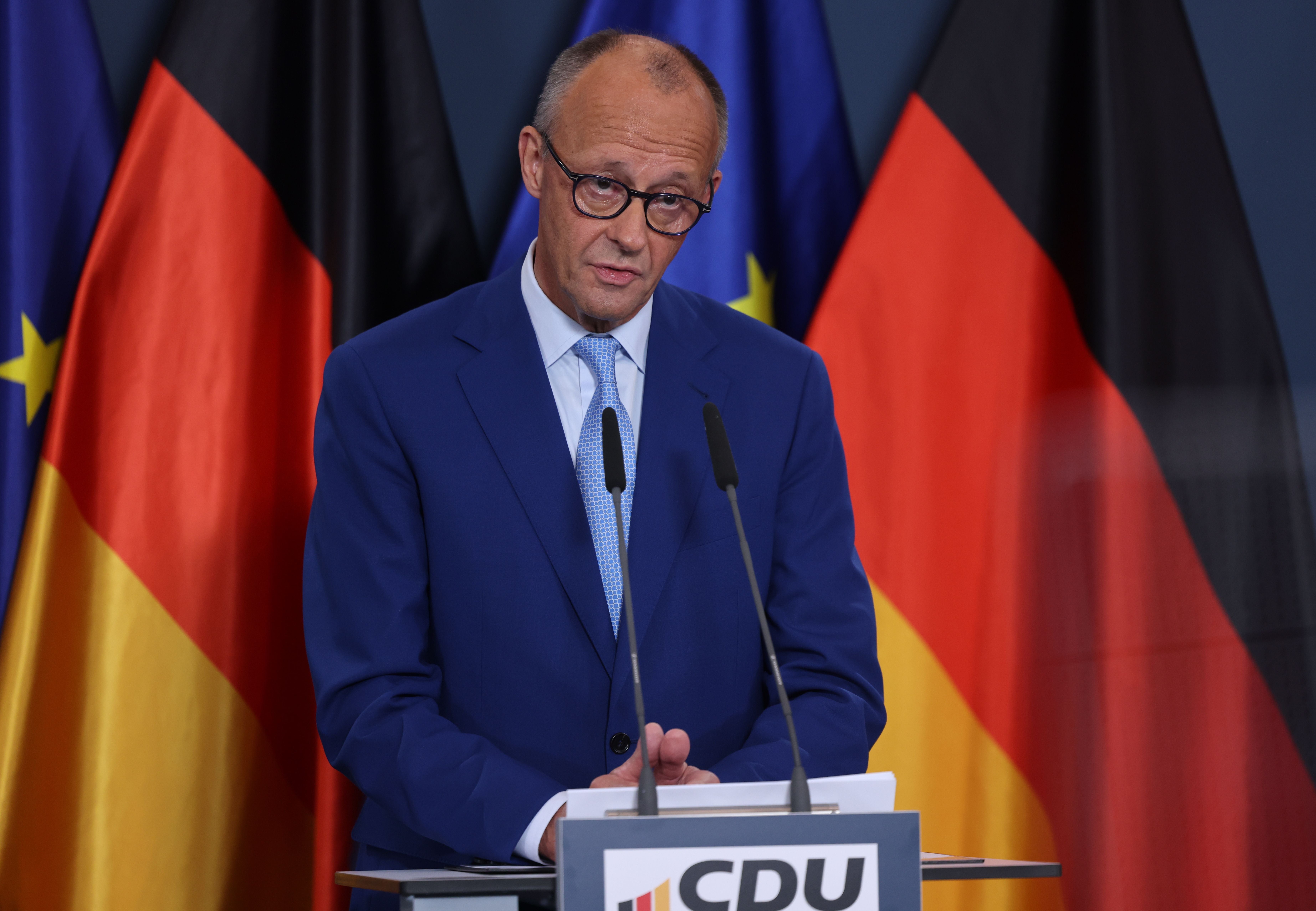 Ein Mann im blauen Anzug spricht bei einer CDU-Pressekonferenz vor deutschen und EU-Flaggen.