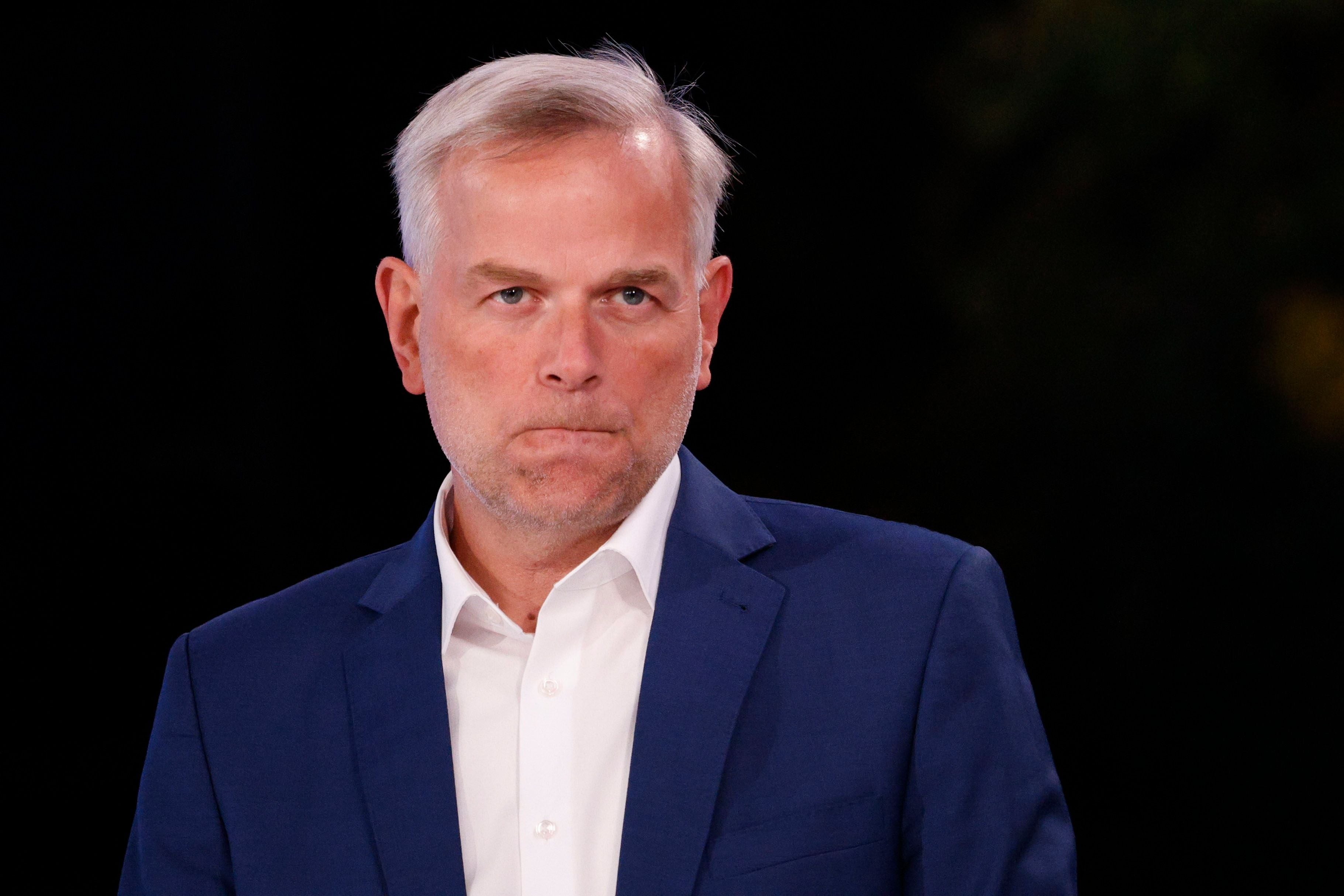 Ein Mann im blauen Anzug schaut ernst in die Kamera, Hintergrund ist dunkel.