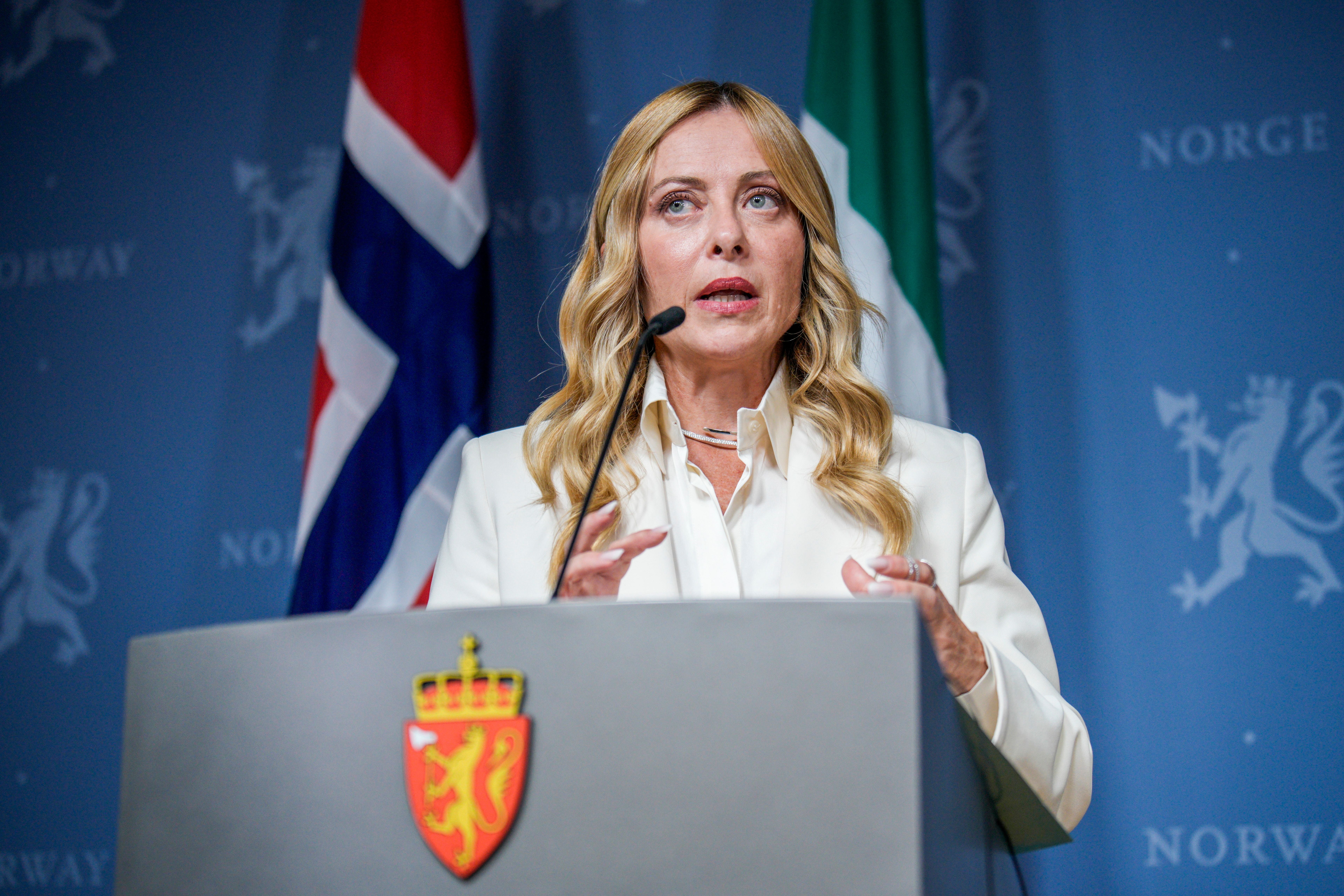 Frau am Rednerpult bei einer Pressekonferenz mit norwegischer und italienischer Flagge im Hintergrund.