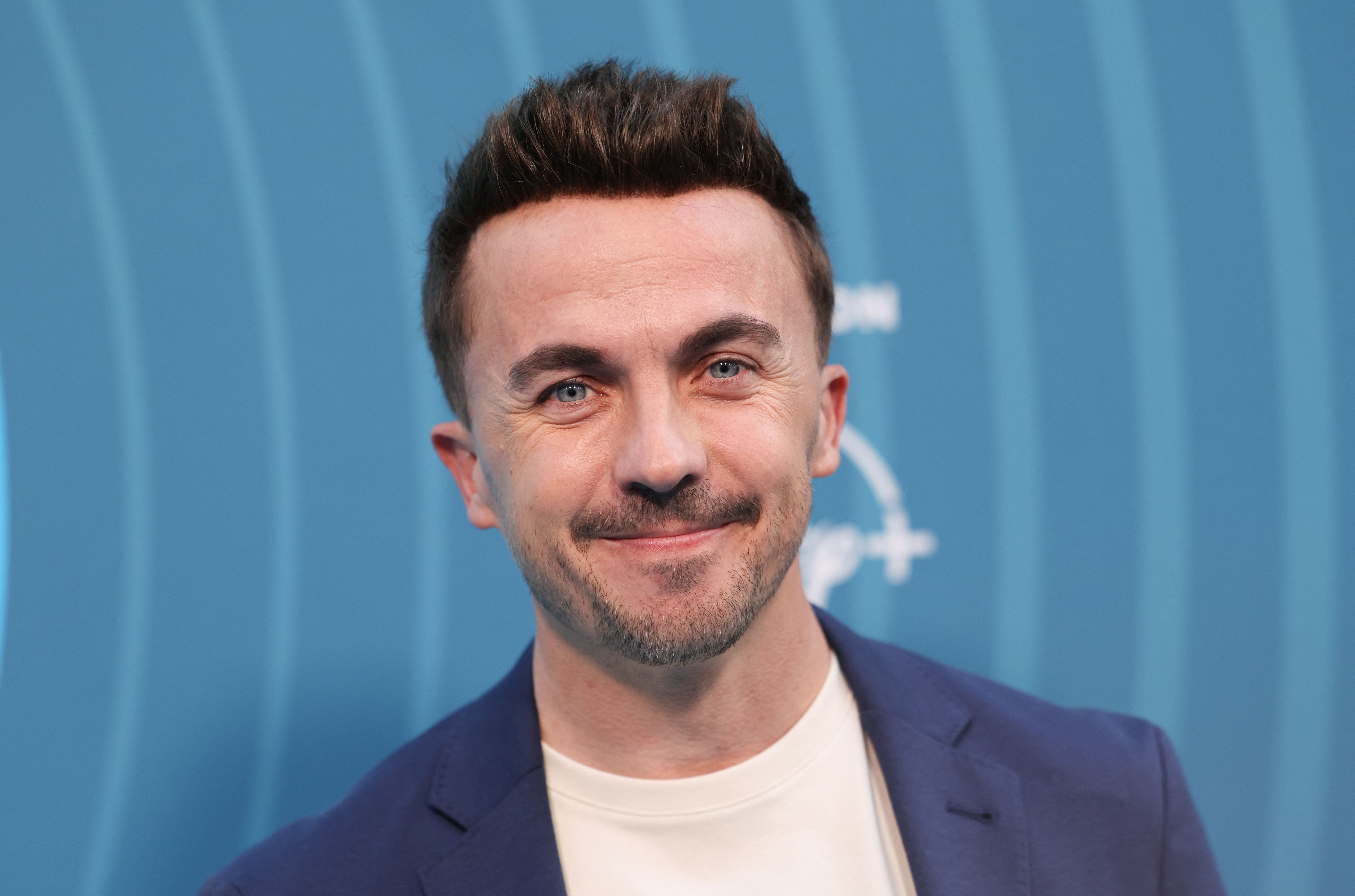 Ein Mann mit kurzem braunem Haar lächelt vor blauem Hintergrund in die Kamera.