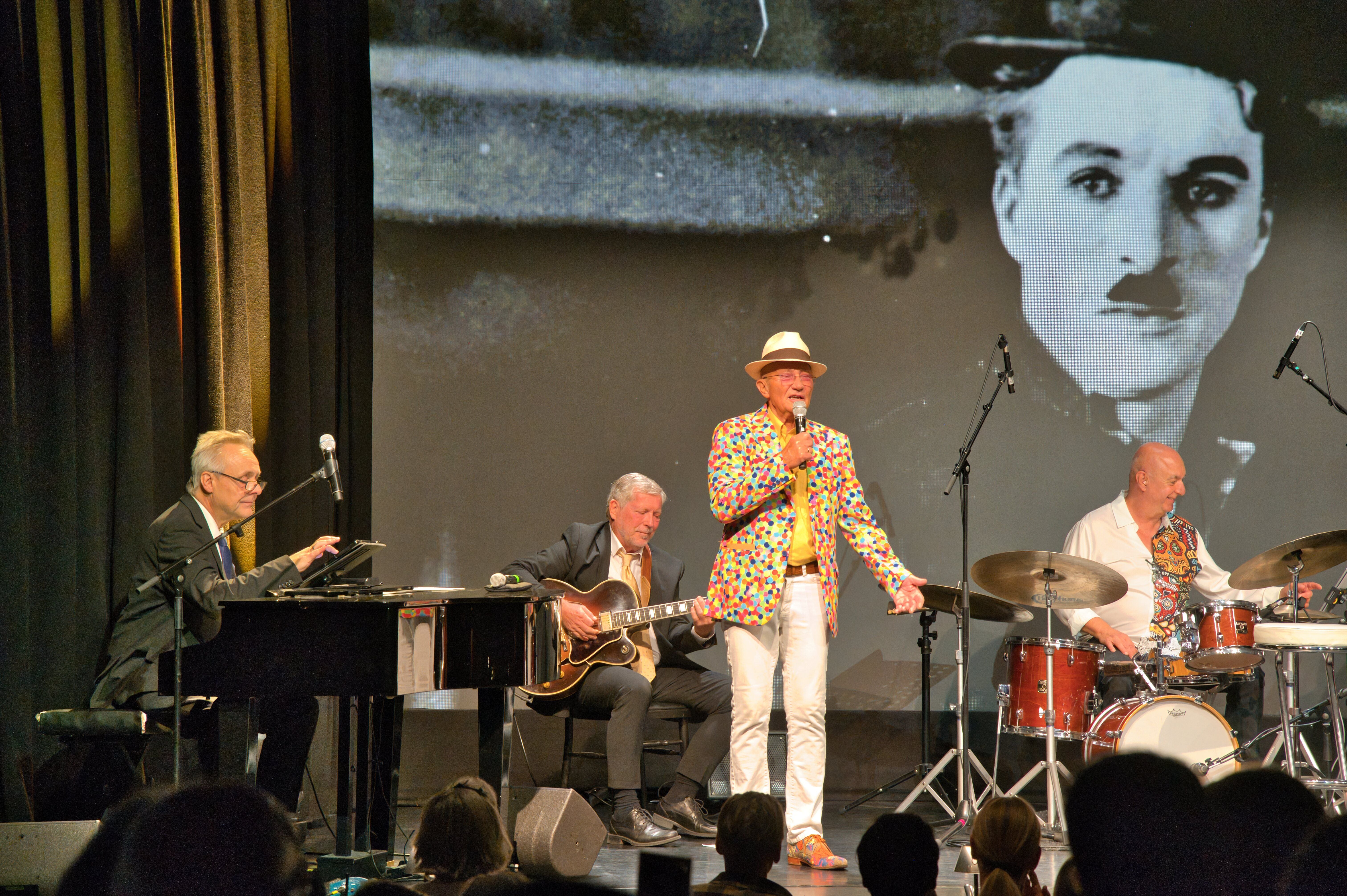 Vier Musiker treten auf einer Bühne auf; ein Mann singt, im Hintergrund ein Foto von Charlie Chaplin.