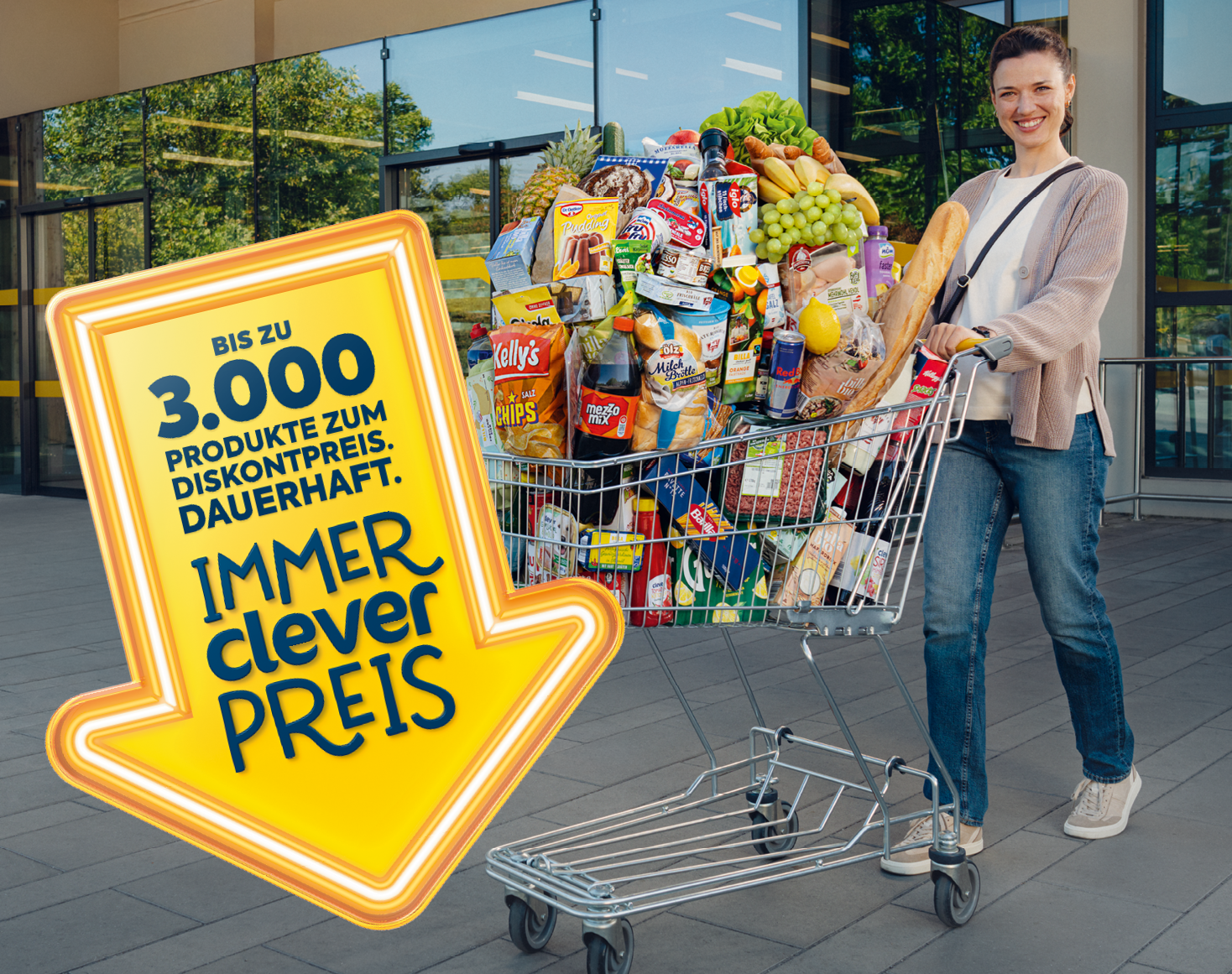 Eingang eines Billa-Supermarkts mit zwei Menschen, Einkaufwagen und Bergen im Hintergrund.