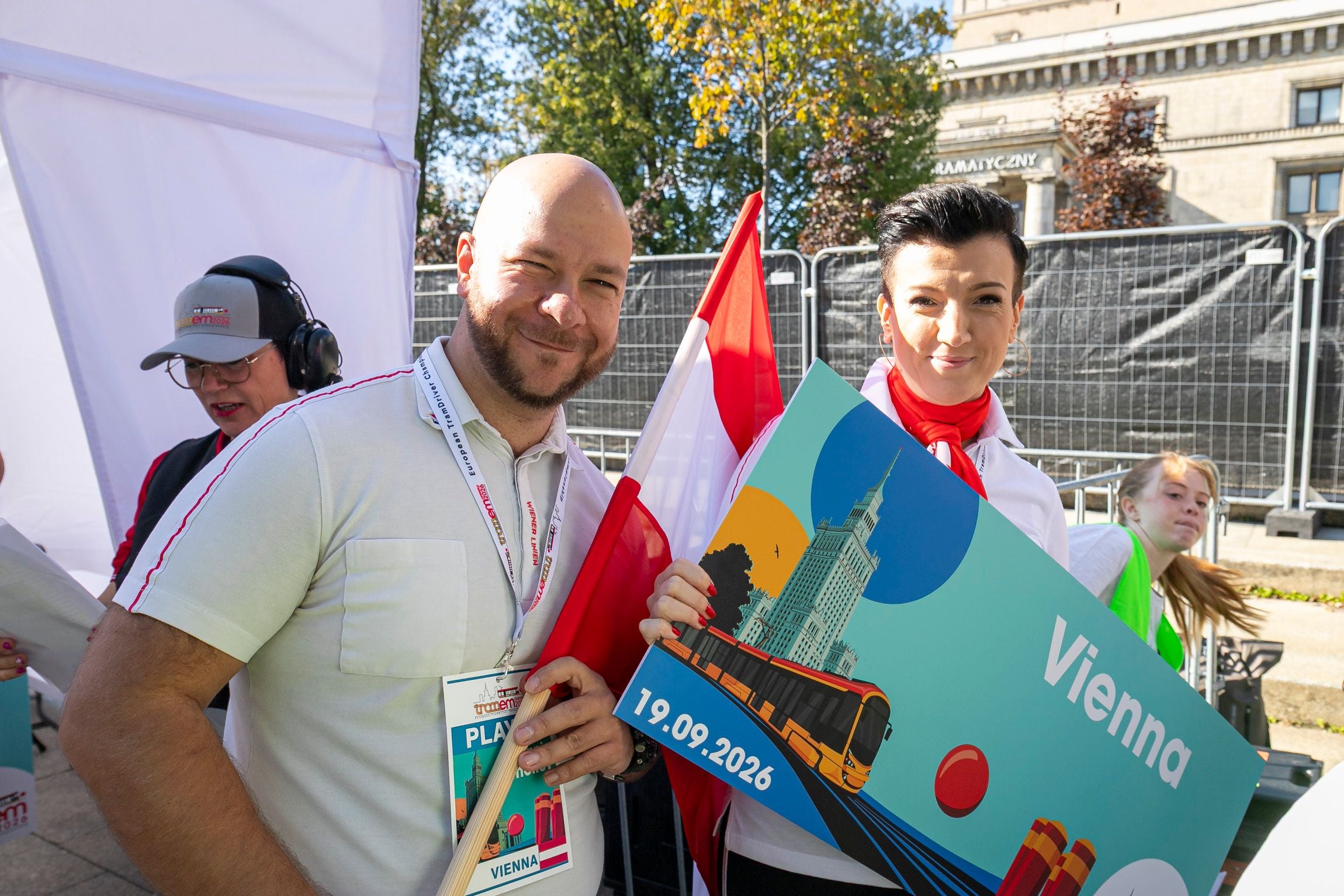 Zwei Personen halten eine Wien-Tafel und eine Fahne bei einer Outdoor-Veranstaltung.