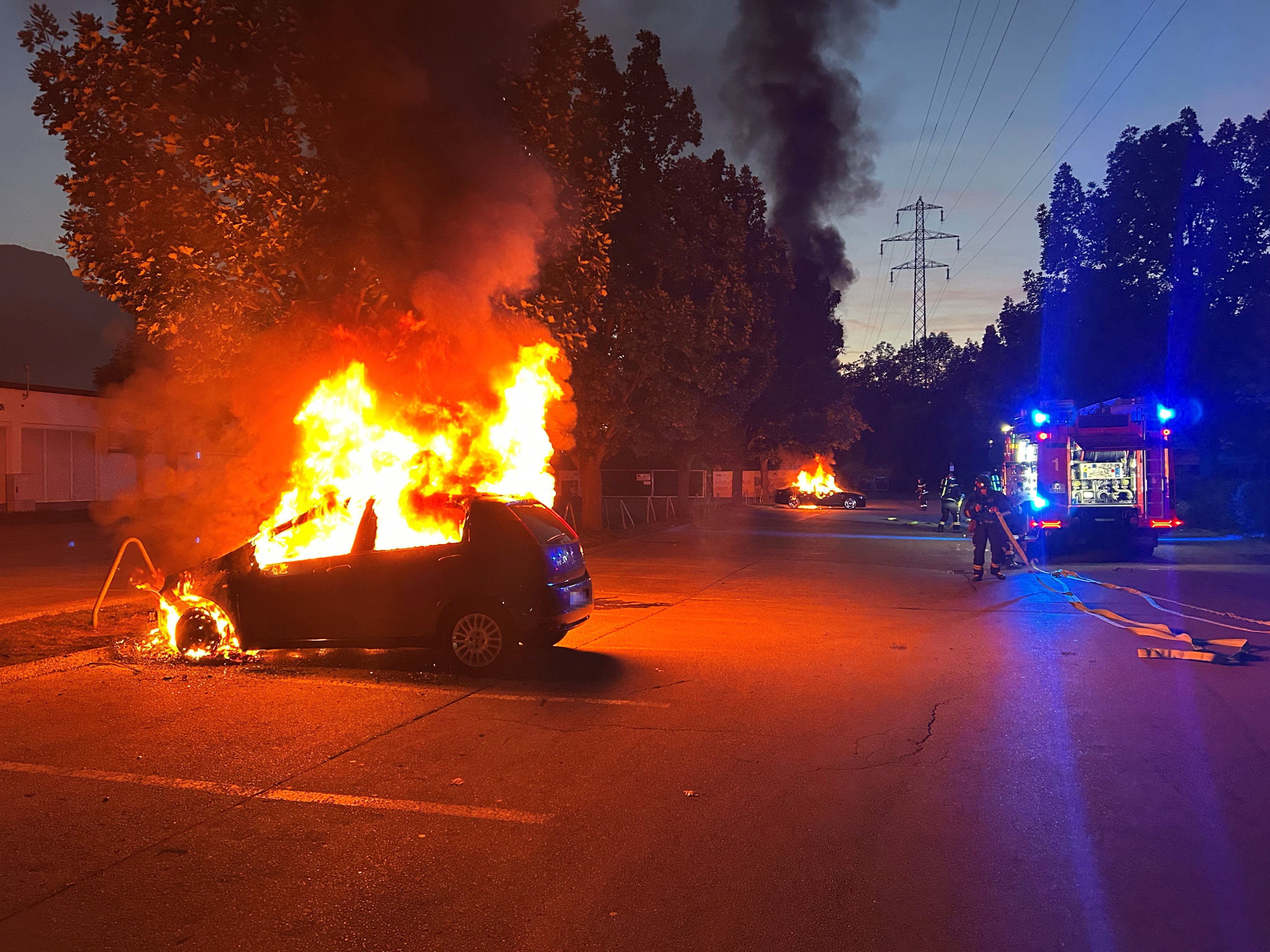 Zwei Autos stehen nachts auf einem Parkplatz in Innsbruck in Flammen, Feuerwehr ist im Einsatz.