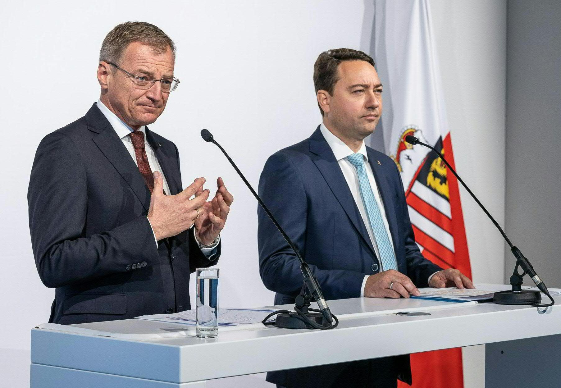 Zwei Männer im Anzug stehen bei einer Pressekonferenz an einem Redepult mit Mikrofonen und Fahne.