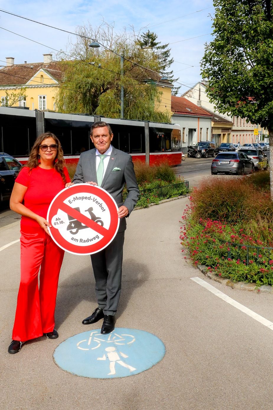 Eine Frau und ein Mann halten ein Schild „E-Moped-Verbot am Radweg“ auf einem Radweg.