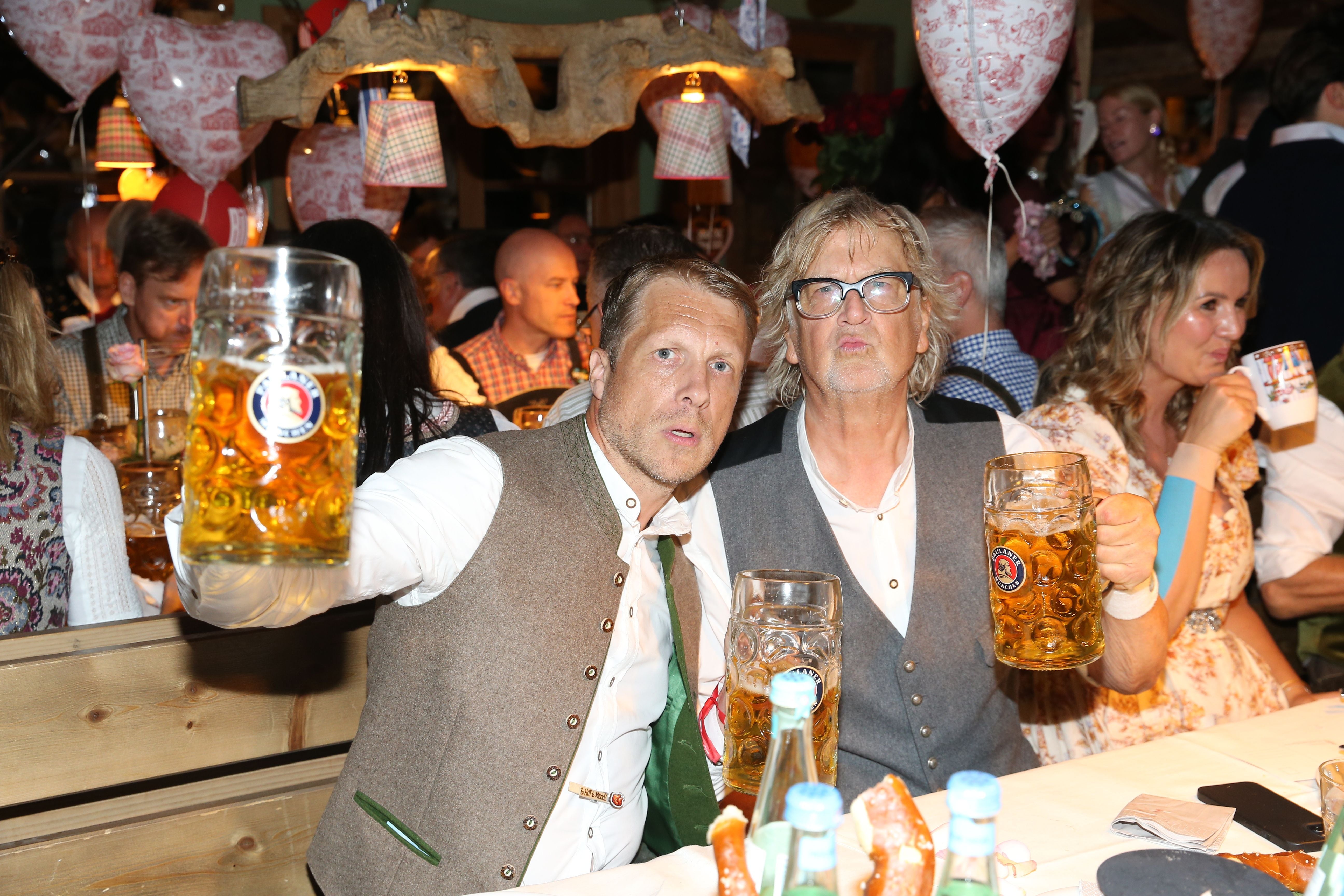 Zwei Männer stoßen mit großen Bierkrügen auf dem Oktoberfest an, im Hintergrund feiern weitere Gäste.