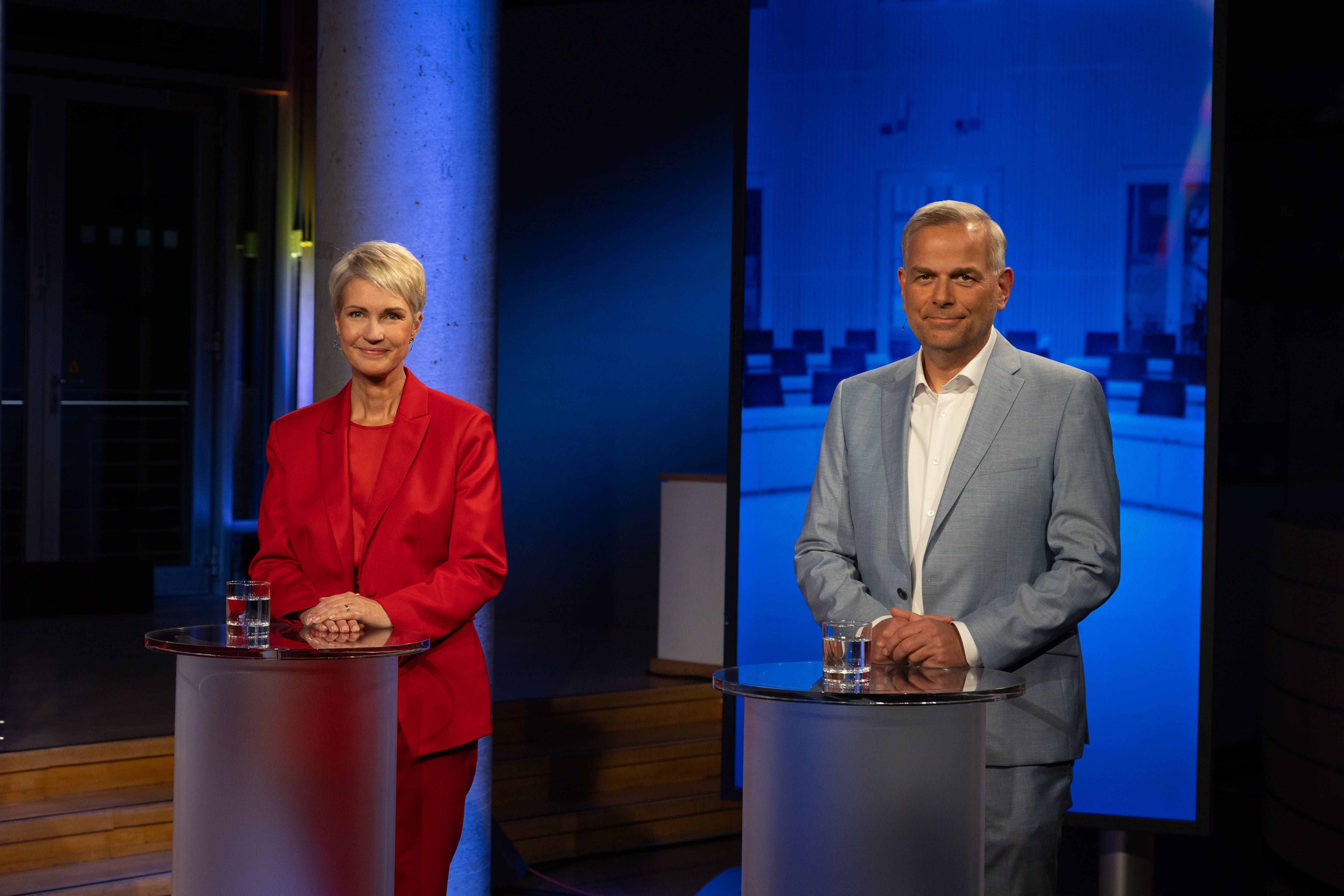 Zwei Personen stehen an Pulten bei einer TV-Debatte mit blauem Hintergrund und Wassergläsern.