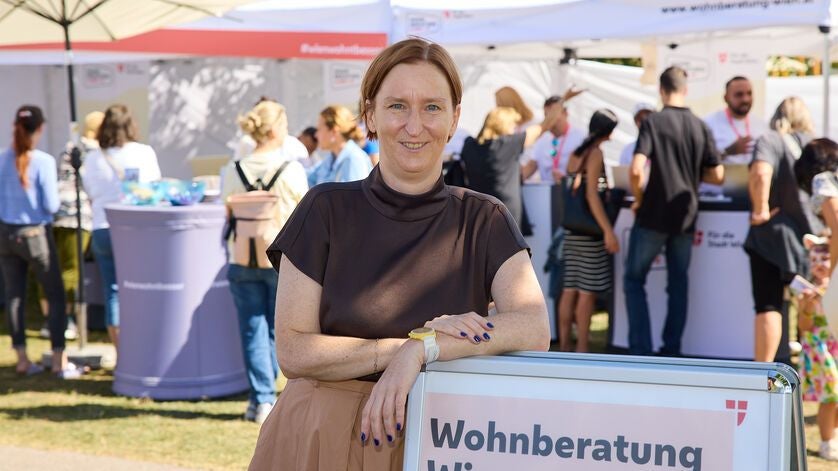 Eine Frau steht lächelnd vor einem Wohnberatung-Stand auf einer Freiluftveranstaltung.