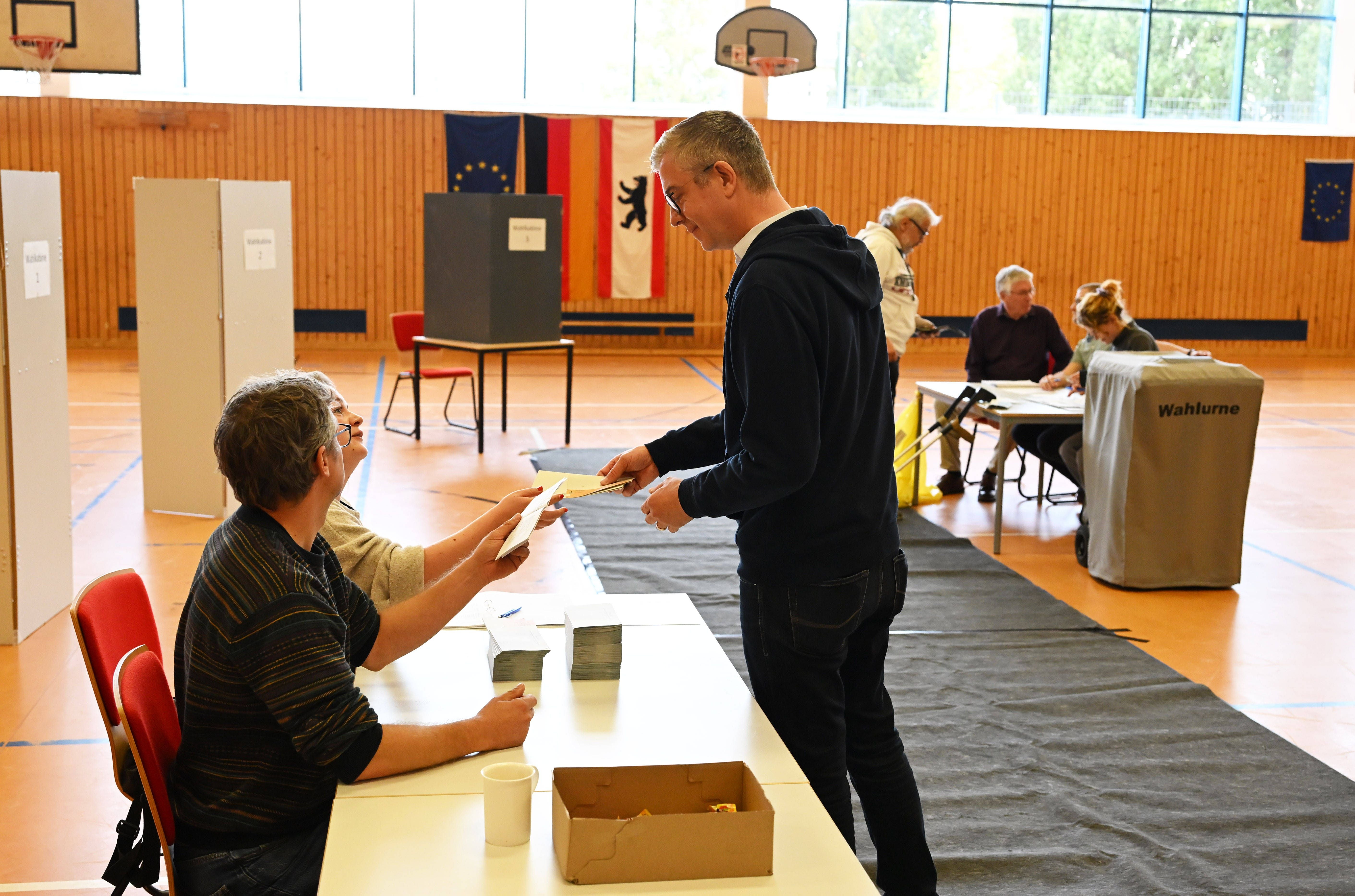 Wähler gibt seinen Stimmzettel an einem Tisch in einem Wahllokal ab, weitere Personen im Hintergrund.