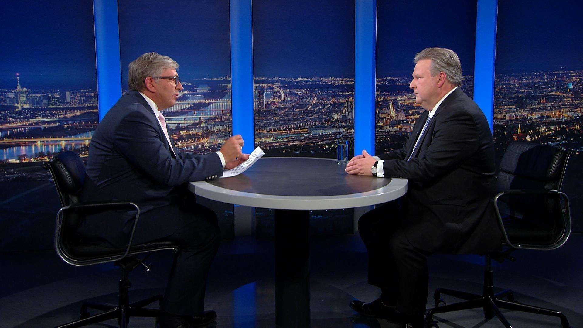 Zwei Männer im Anzug führen ein Fernsehinterview an einem runden Tisch vor Stadtpanorama.