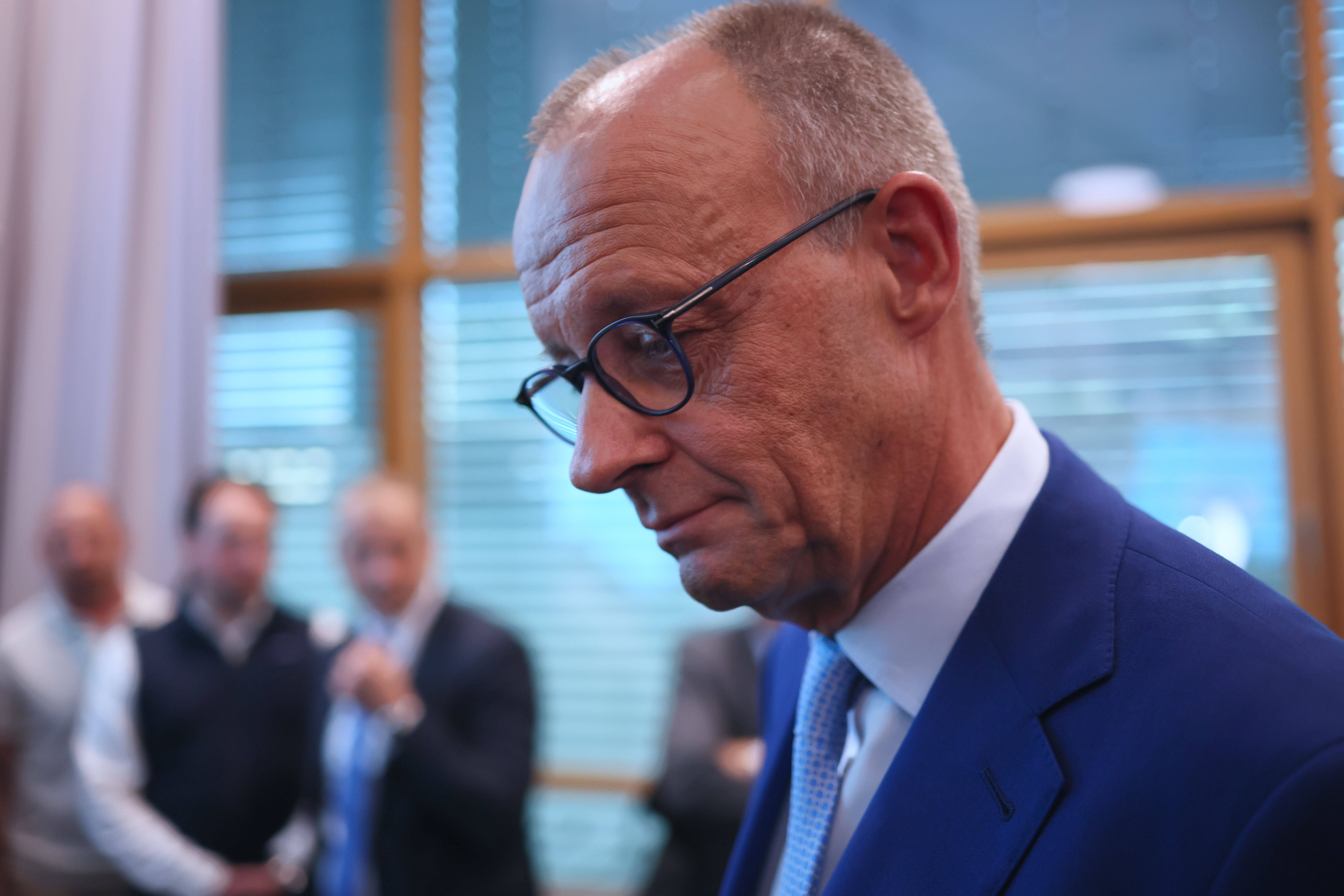 Ein Mann mit Brille im blauen Anzug steht nachdenklich in einem Raum, unscharfe Personen im Hintergrund.