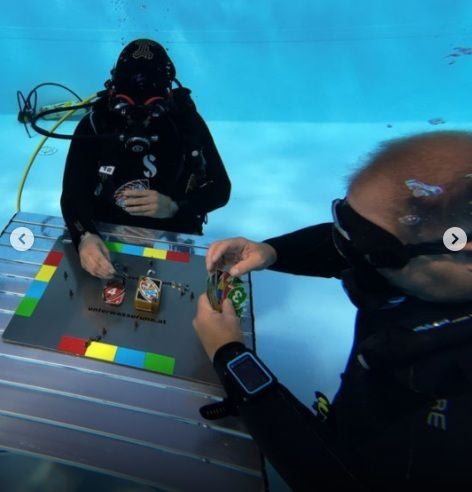 Zwei Taucher spielen UNO an einem Tisch unter Wasser.