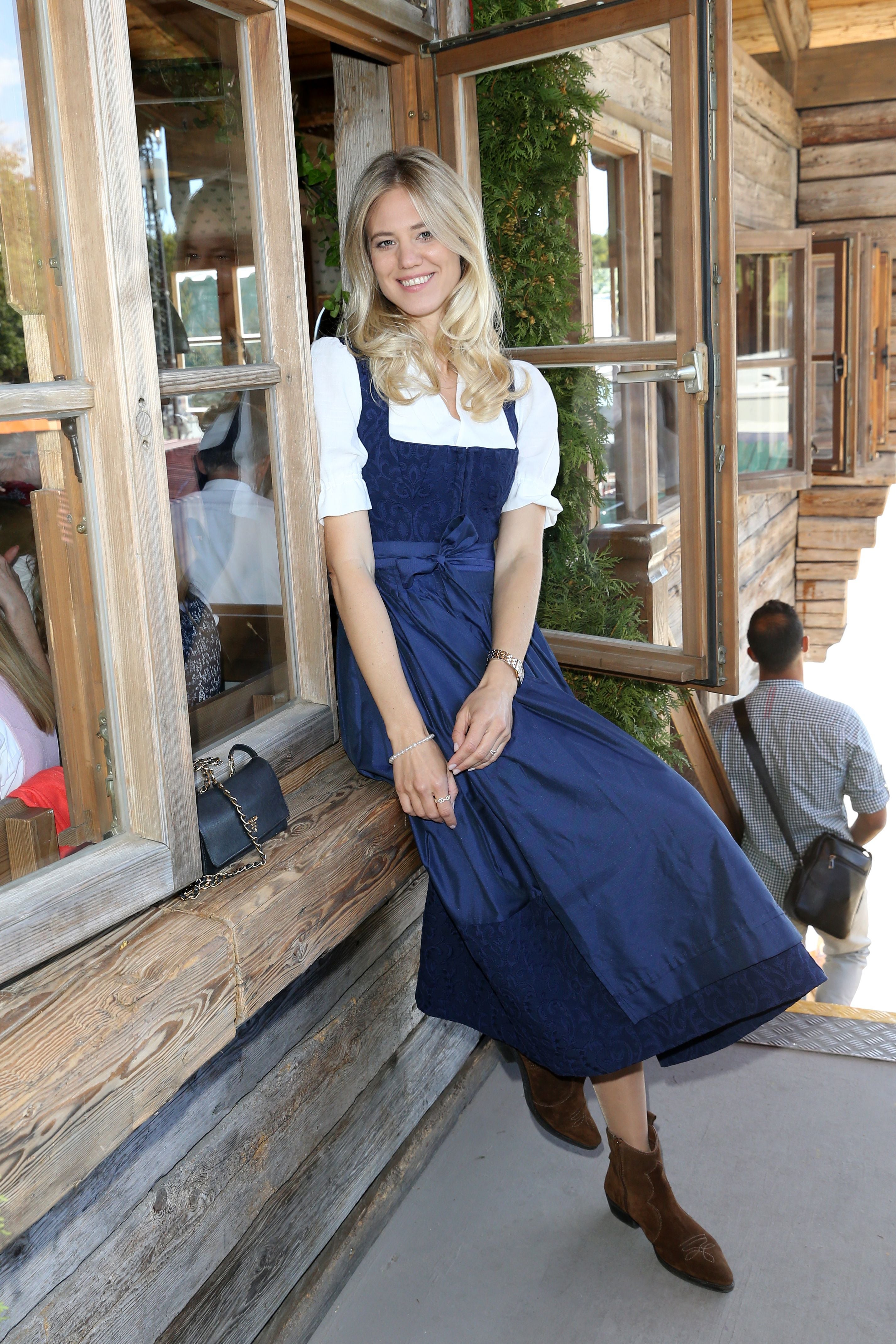 Frau in blauem Dirndl sitzt lächelnd auf einer Holzbank vor einem Fenster eines traditionellen Hauses.